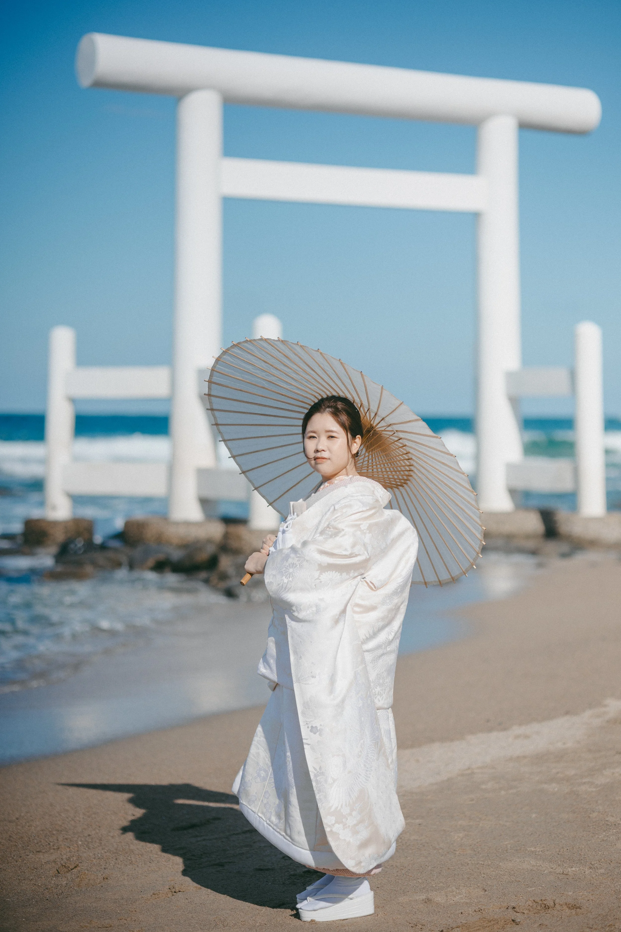 A woman in traditional Japanese attire holding a parasol on the beach with a large white Torii gate in the background.