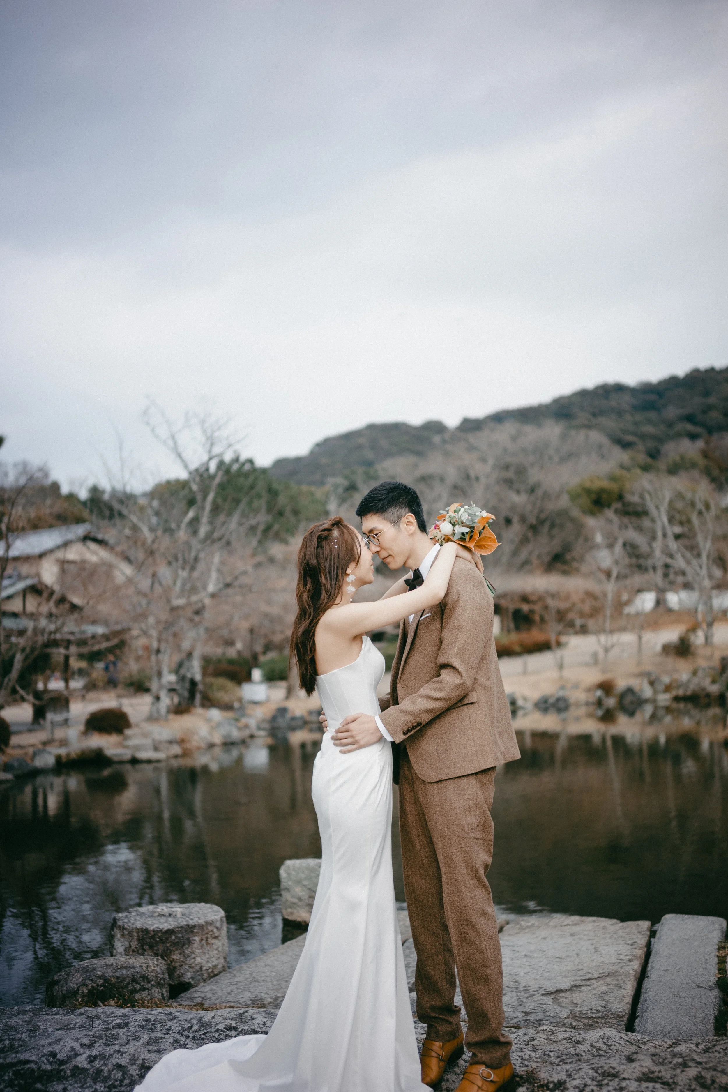 A bride and groom embrace on a natural stone platform near a pond with leafless trees and hills in the background, during a wedding photo shoot.