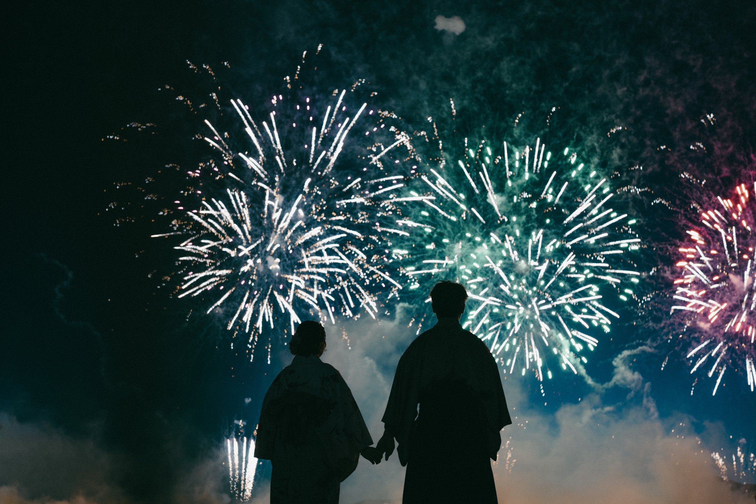 Silhouettes of a couple in traditional Japanese clothing holding hands, watching fireworks in the night sky.