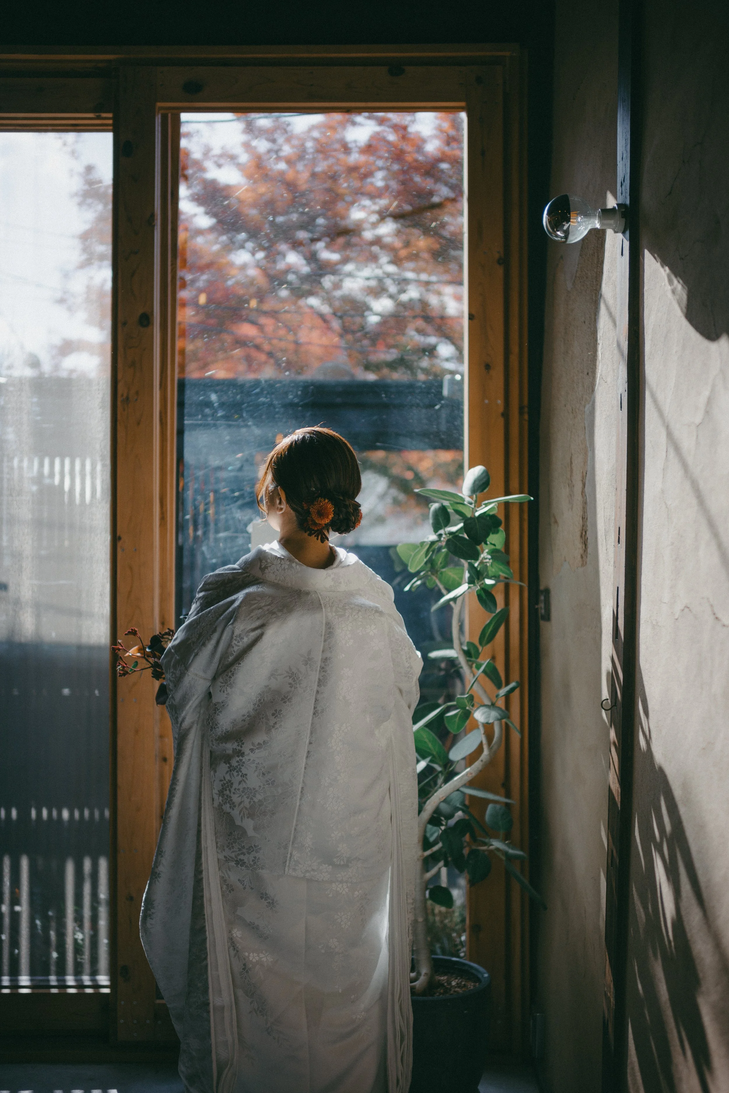 A woman dressed in a traditional kimono standing indoors by a large window with autumn trees outside.