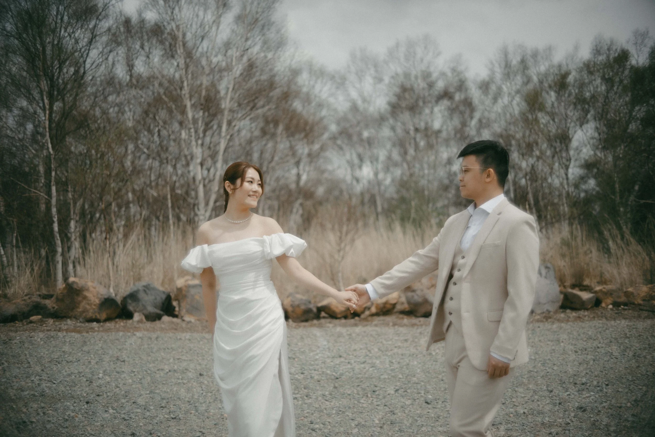 A bride and groom holding hands outdoors on a gravel path with trees in the background, dressed in wedding attire.