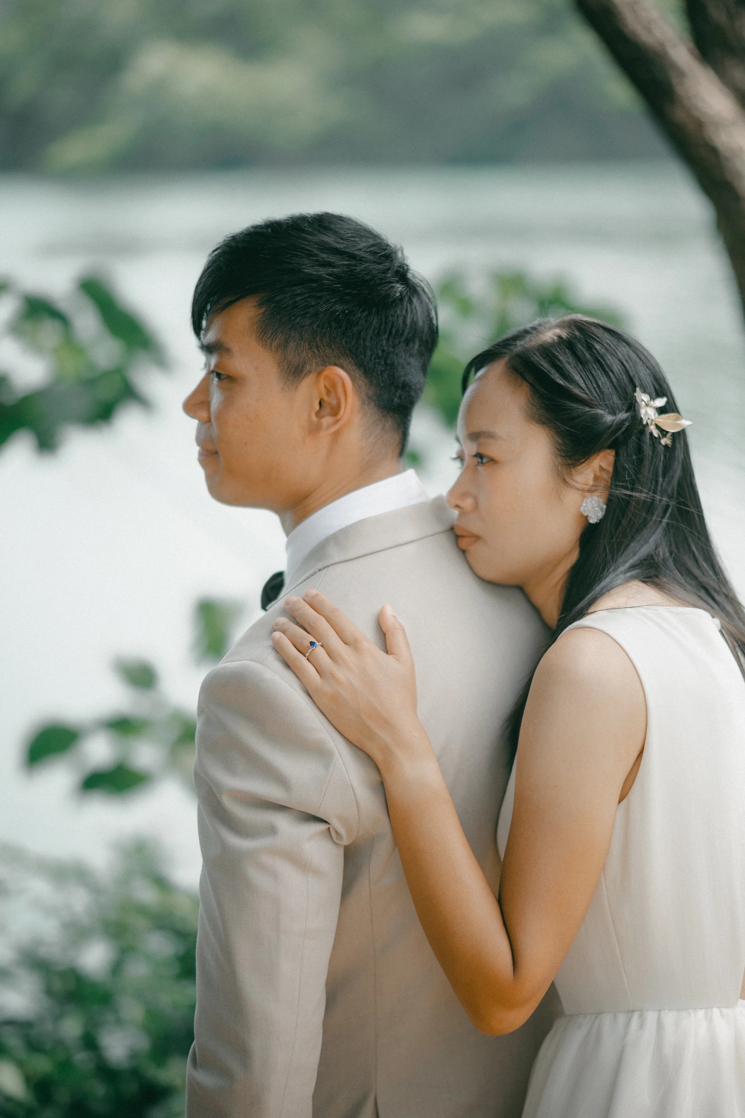 A couple standing close together outdoors, with a lake in the background. The woman rests her head on the man's shoulder, and her hand with a ring is on his shoulder, suggesting a romantic or wedding scene.