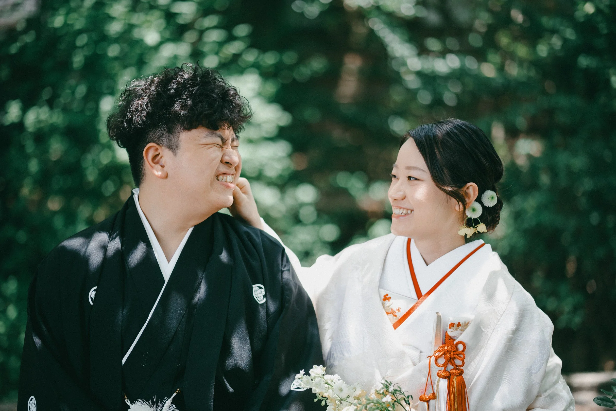 A couple dressed in traditional Japanese wedding attire, with the man in black and the woman in white, smiling and looking at each other outdoors in a natural setting.