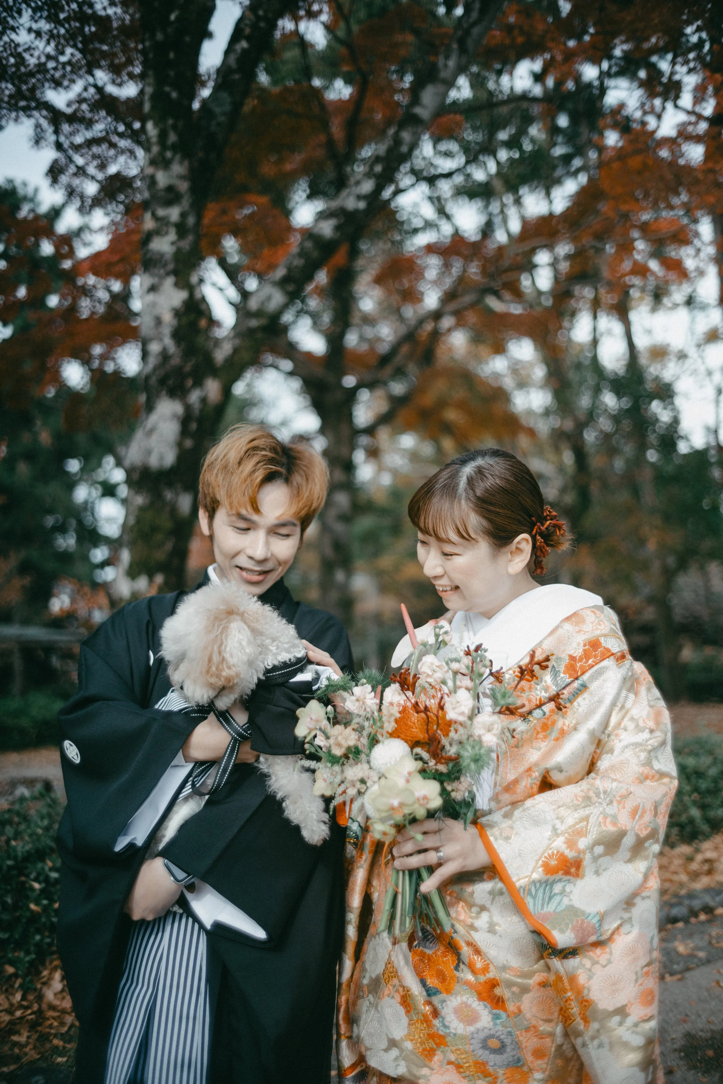A woman in a traditional Japanese kimono holding a bouquet, smiling as she looks at a small, fluffy dog in a jacket held by a man in a kimono outdoors with autumn leaves.
