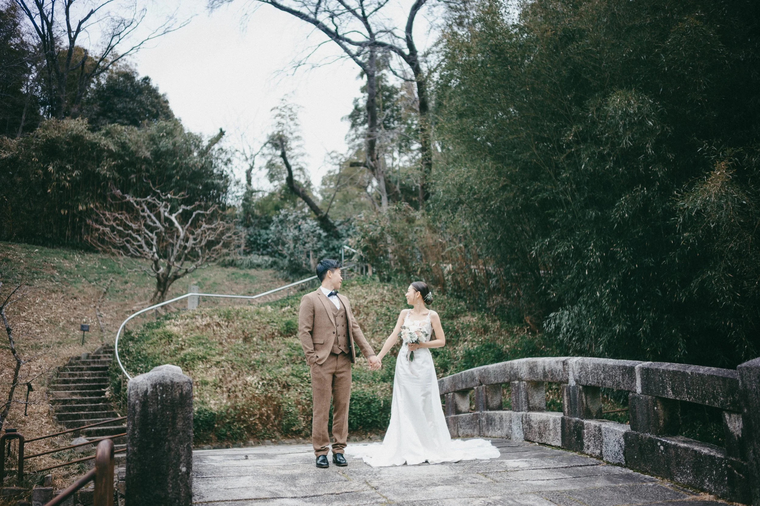 A bride and groom standing on a small stone bridge, holding hands, outdoors in a wooded area during daytime.