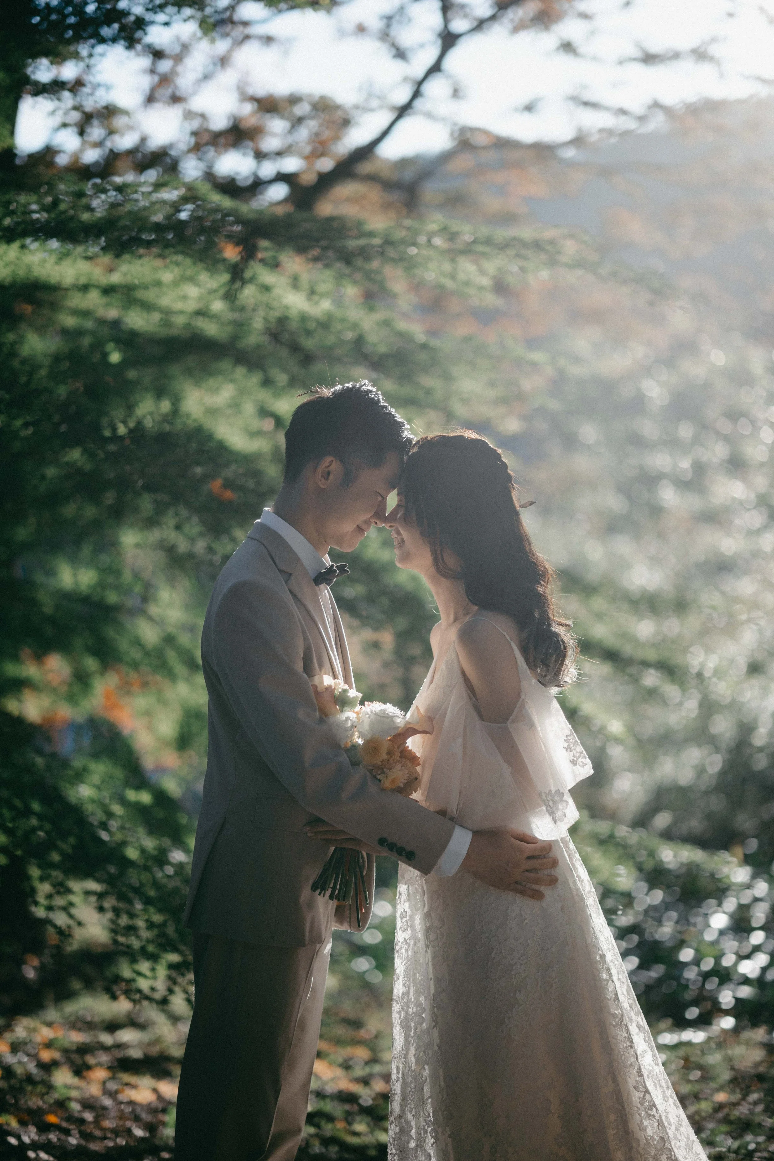 A bride and groom standing close, touching foreheads in a wooded outdoor setting, with sunlight filtering through trees in the background.