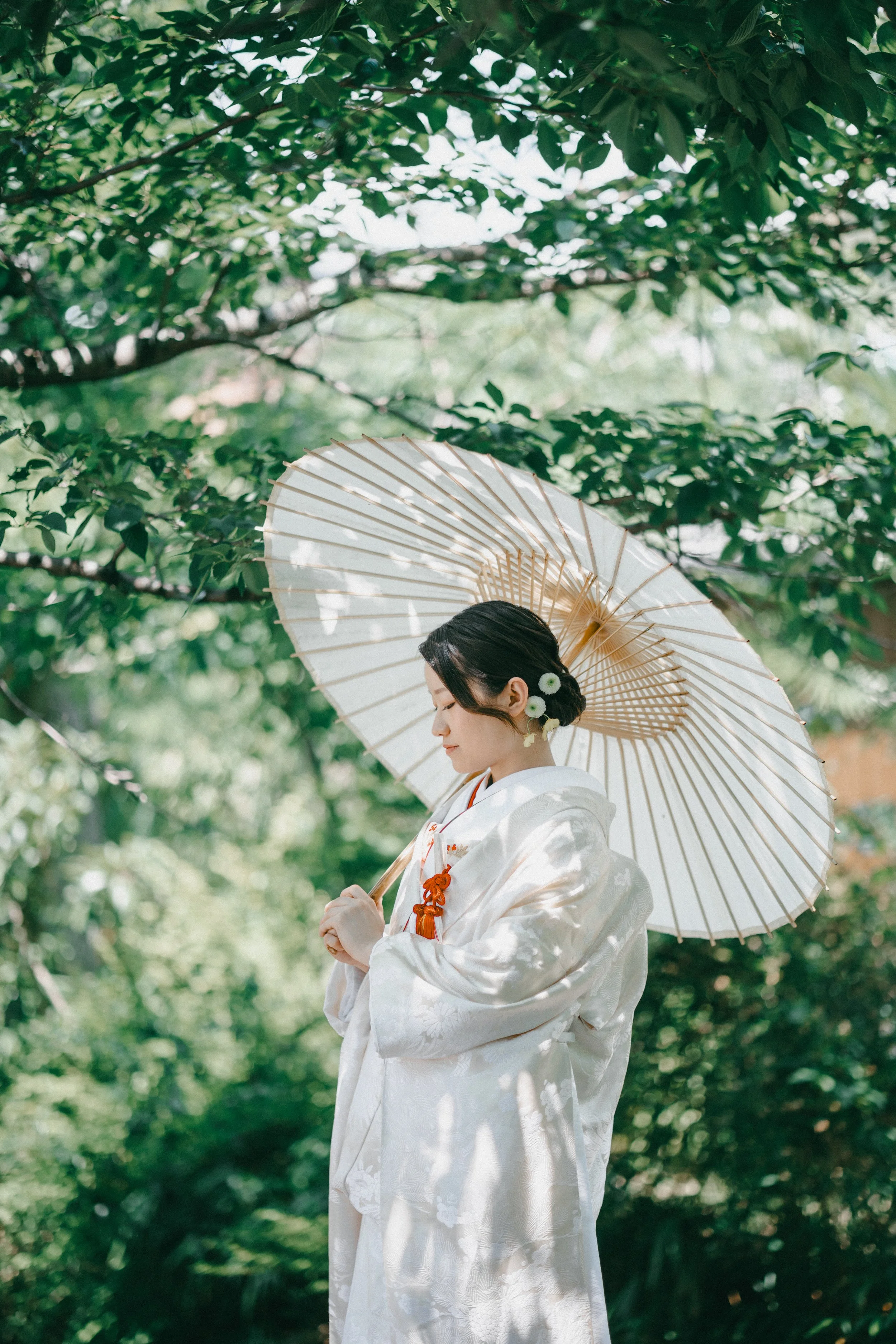 A woman dressed in a traditional white kimono holding a paper parasol, standing in a lush green outdoor setting with trees.