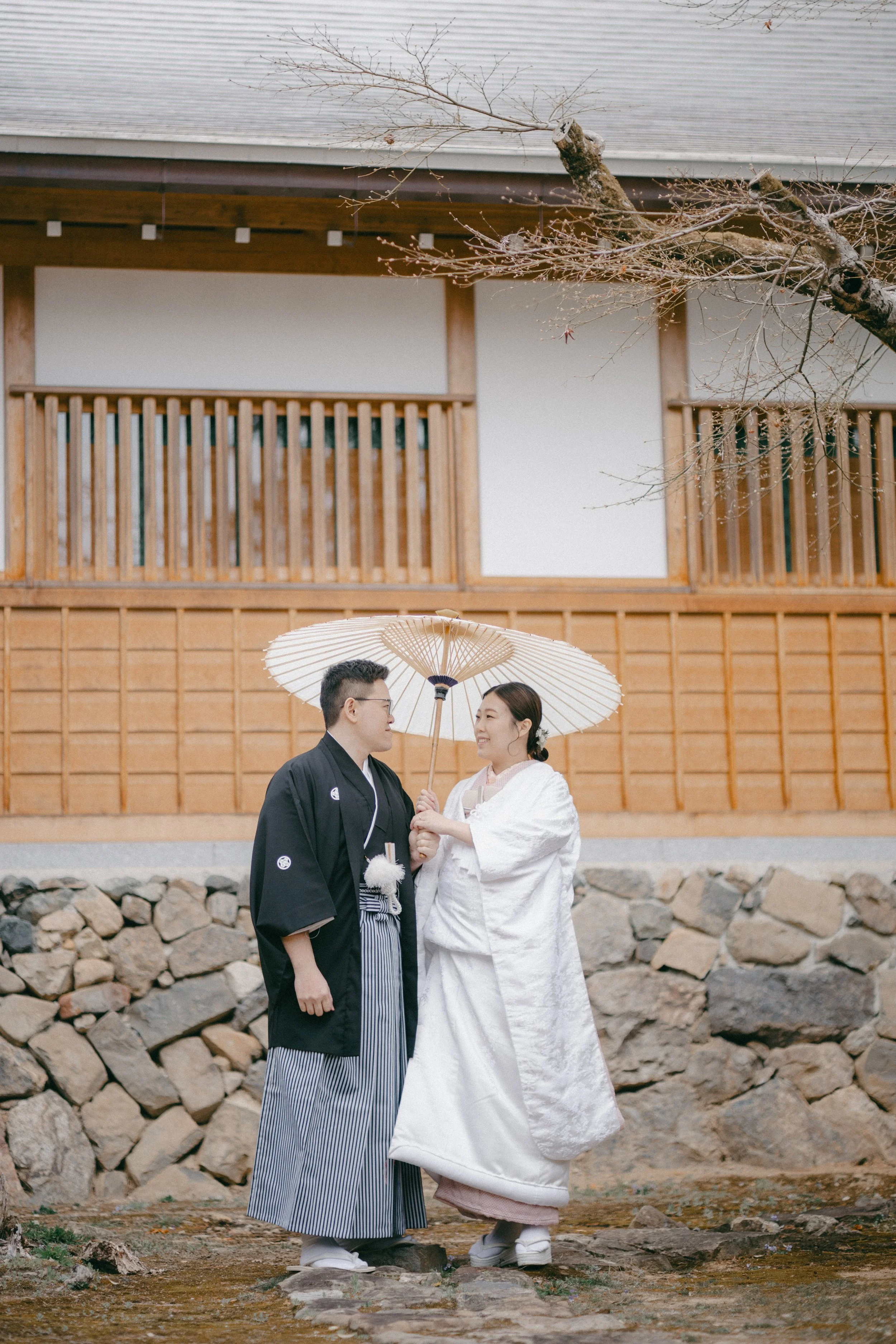 A couple dressed in traditional Japanese wedding attire standing outdoors on a stone path, sharing an umbrella. The man wears a black kimono with white patterns, and the woman wears a white wedding kimono, holding a paper parasol.