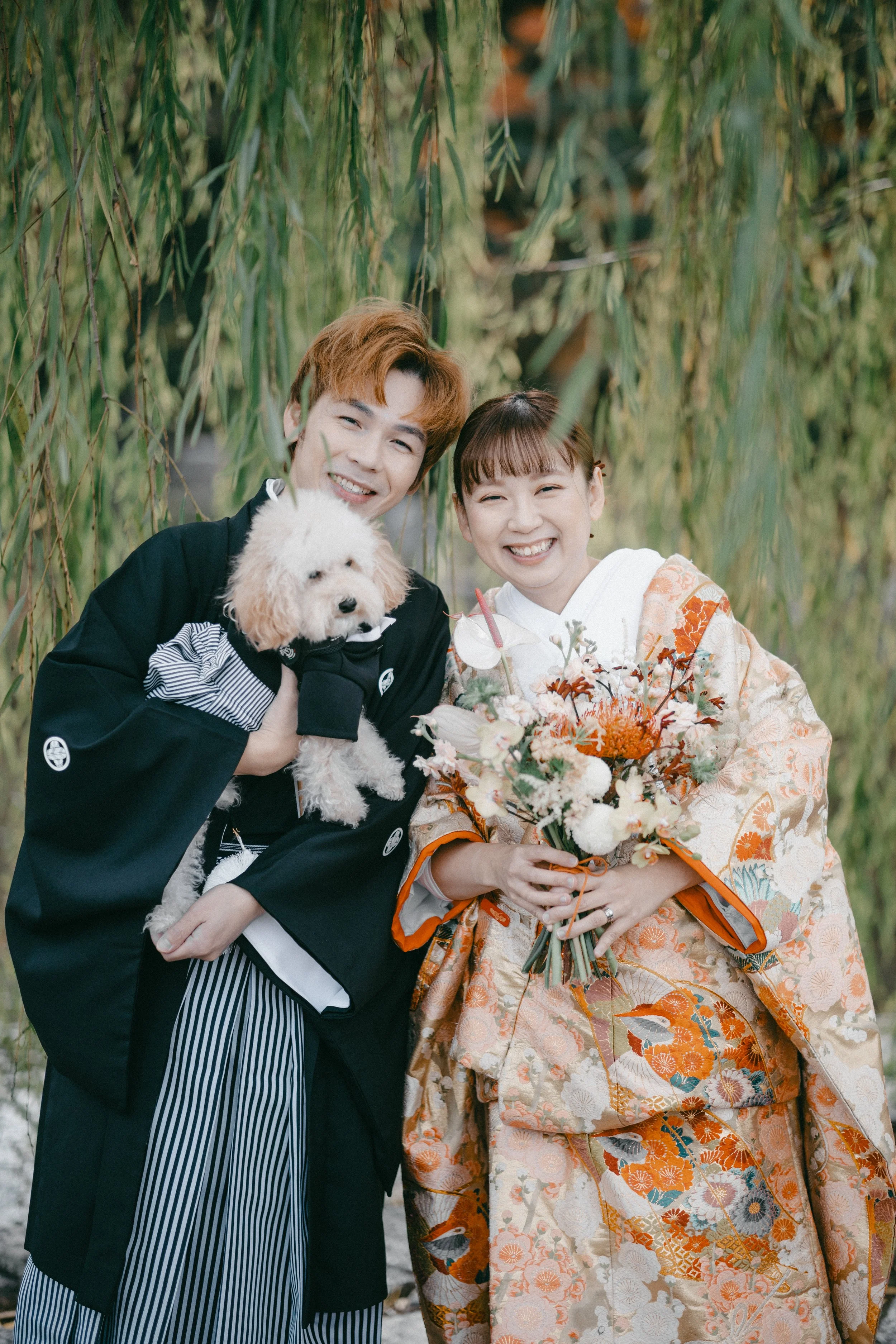 A smiling couple, dressed in traditional Japanese clothing, posing with a woman holding a bouquet of flowers and a man holding a small white dog, standing under a willow tree.