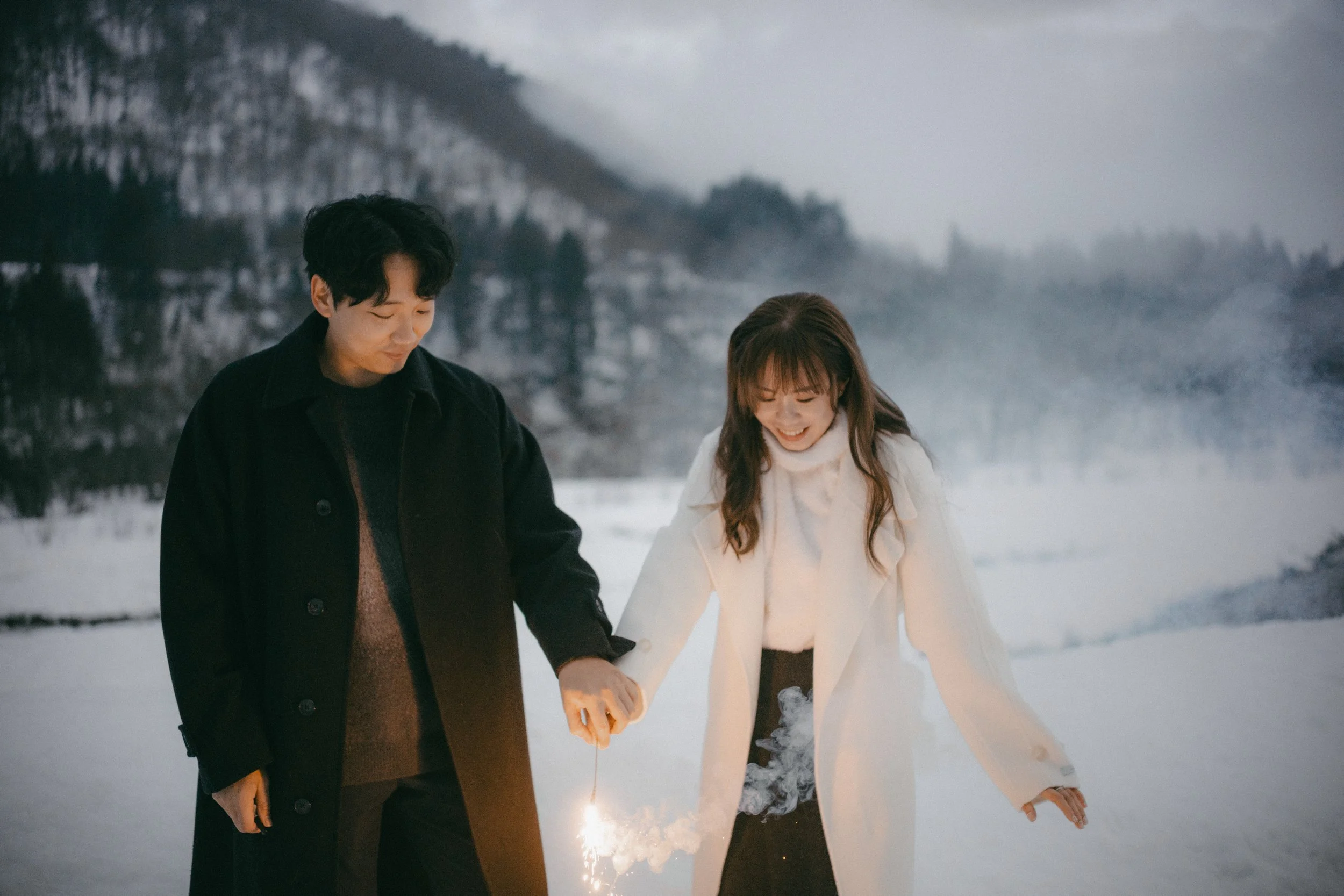A man and woman holding sparklers in snowy outdoor landscape during winter, smiling and enjoying the moment.
