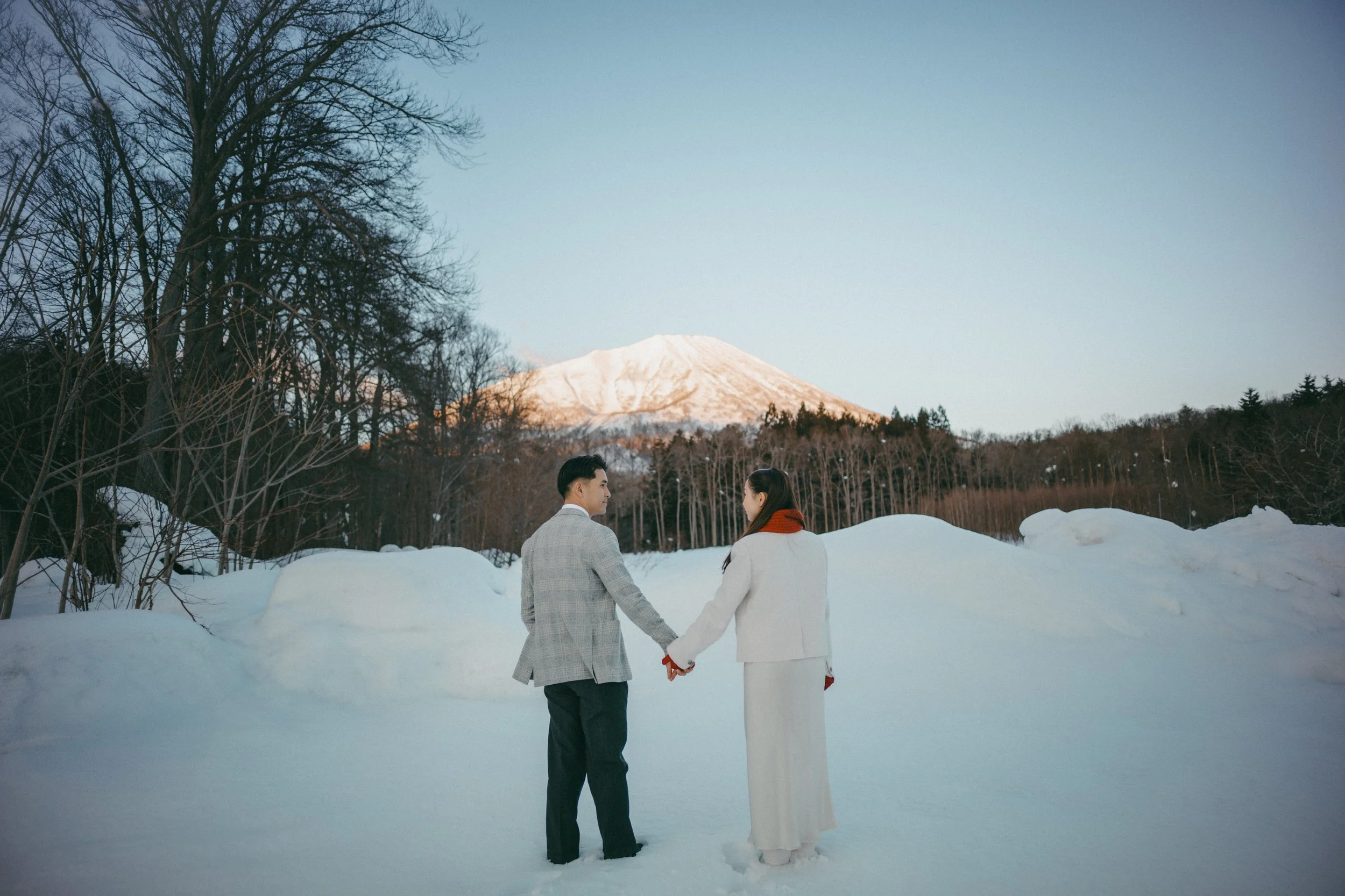 A couple holding hands in a snowy landscape with Mount Fuji in the background, trees, and blue sky.