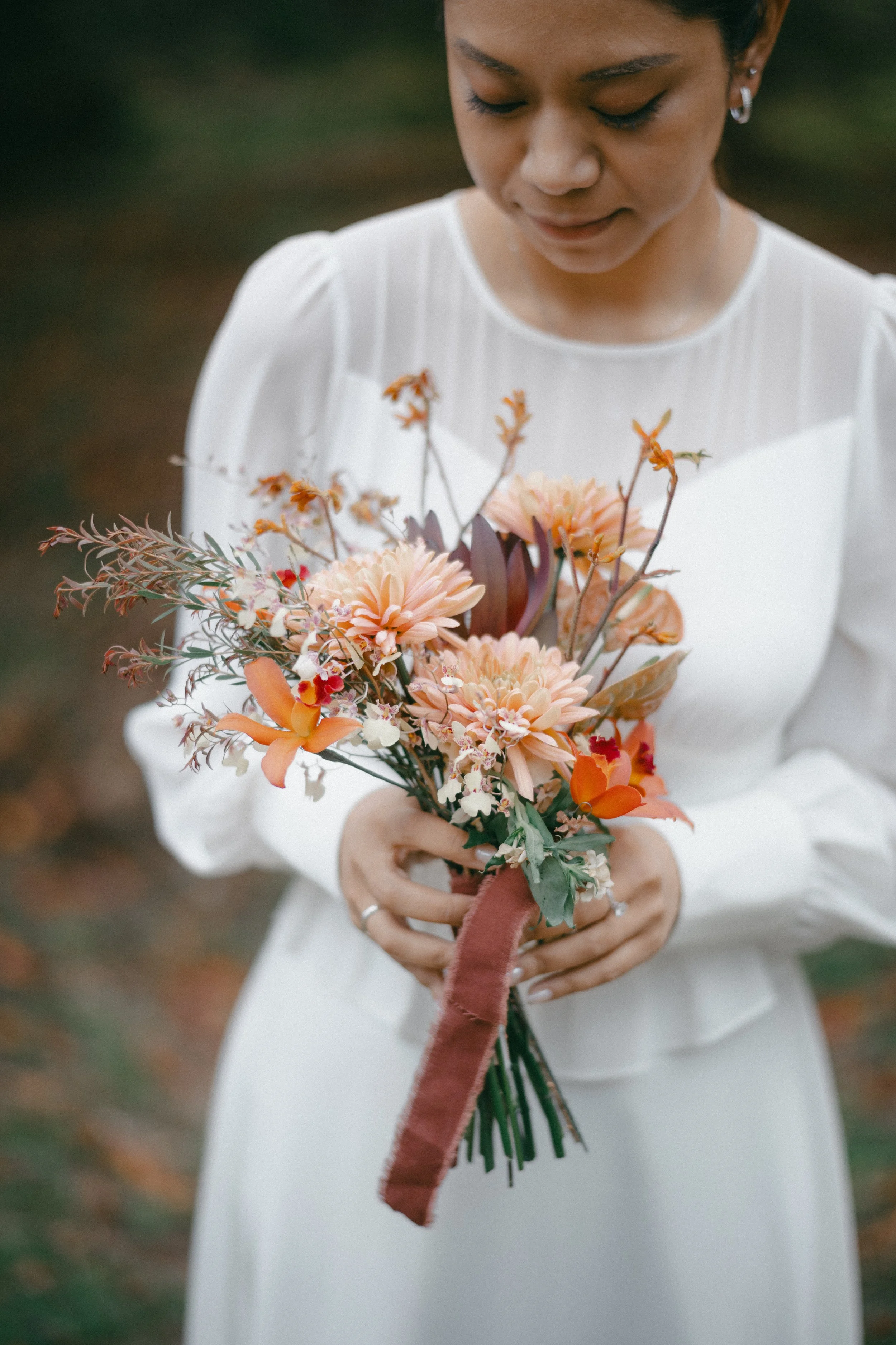 A woman in a white dress holding a bouquet of peach, orange, and cream flowers with greenery and a brown ribbon.