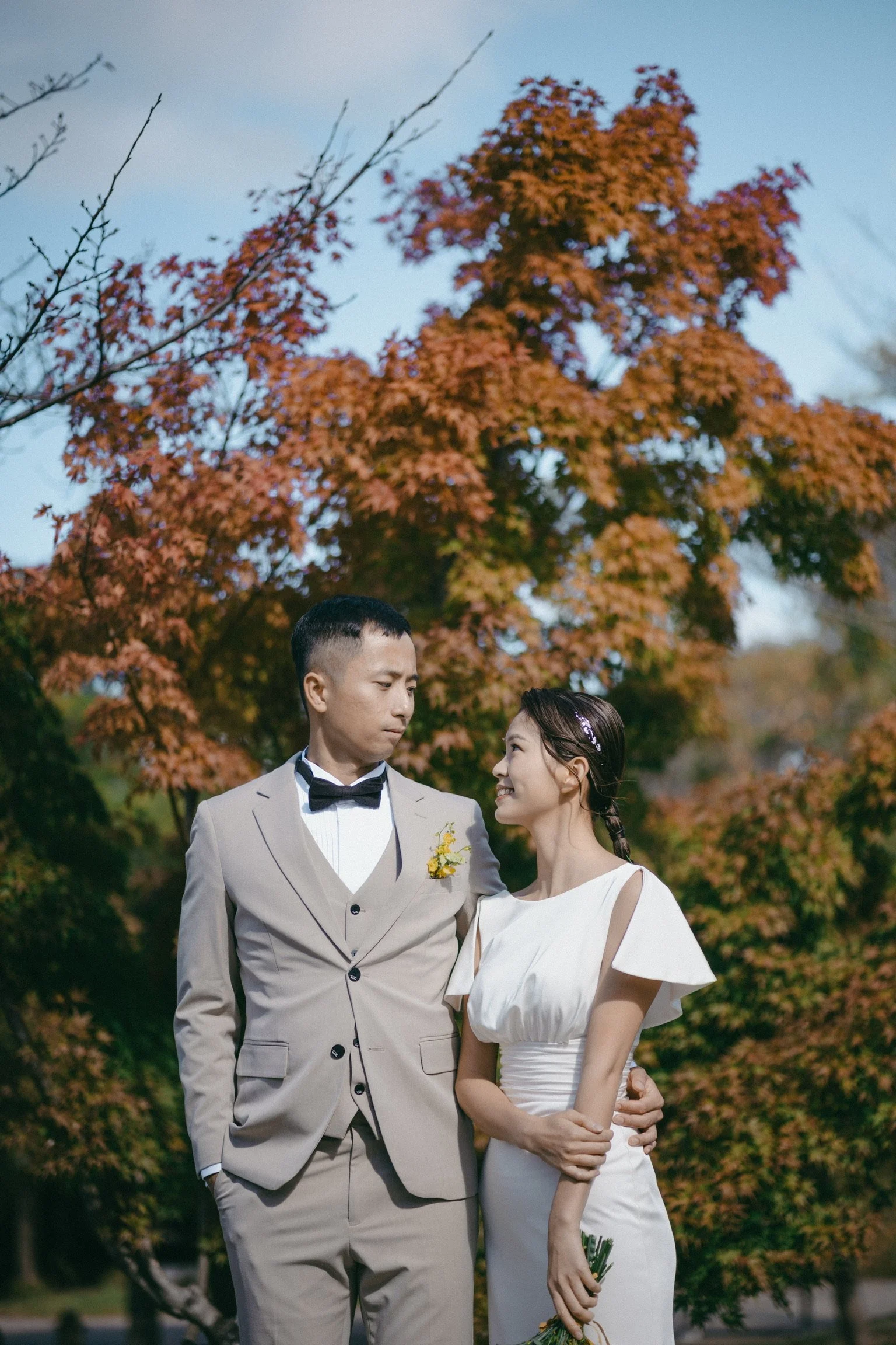 A bride and groom standing outdoors with trees in the background, the bride looking at the groom and smiling.