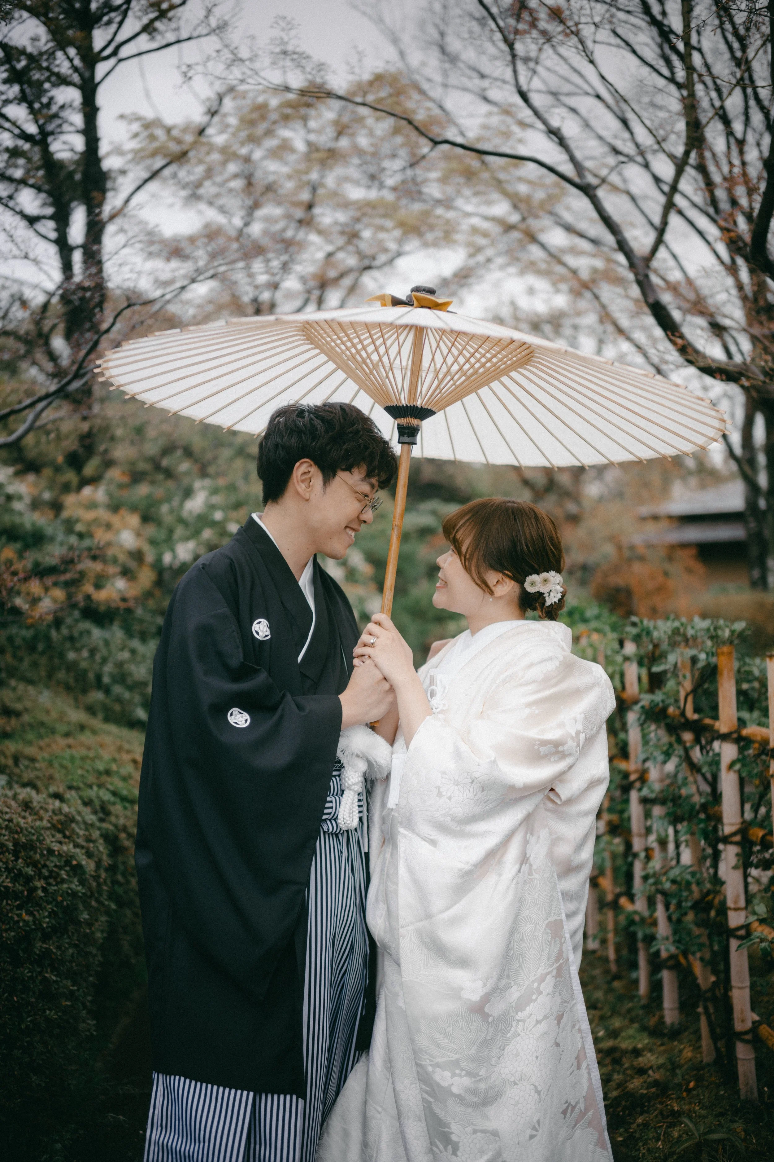 A couple dressed in traditional Japanese wedding attire sharing a moment under a parasol outdoors during autumn.