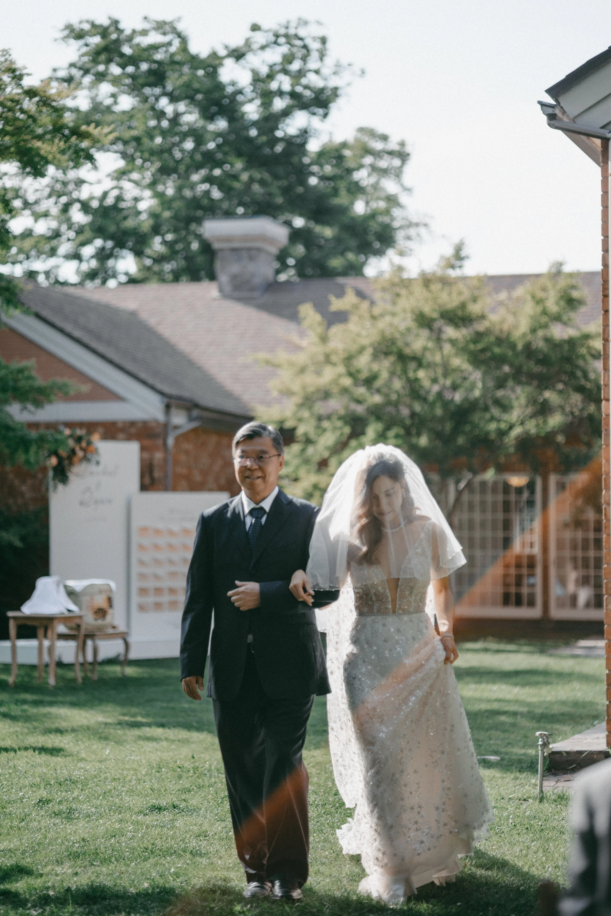 A bride in a wedding dress and veil walking arm-in-arm with a man in a black suit outdoors in sunlight.
