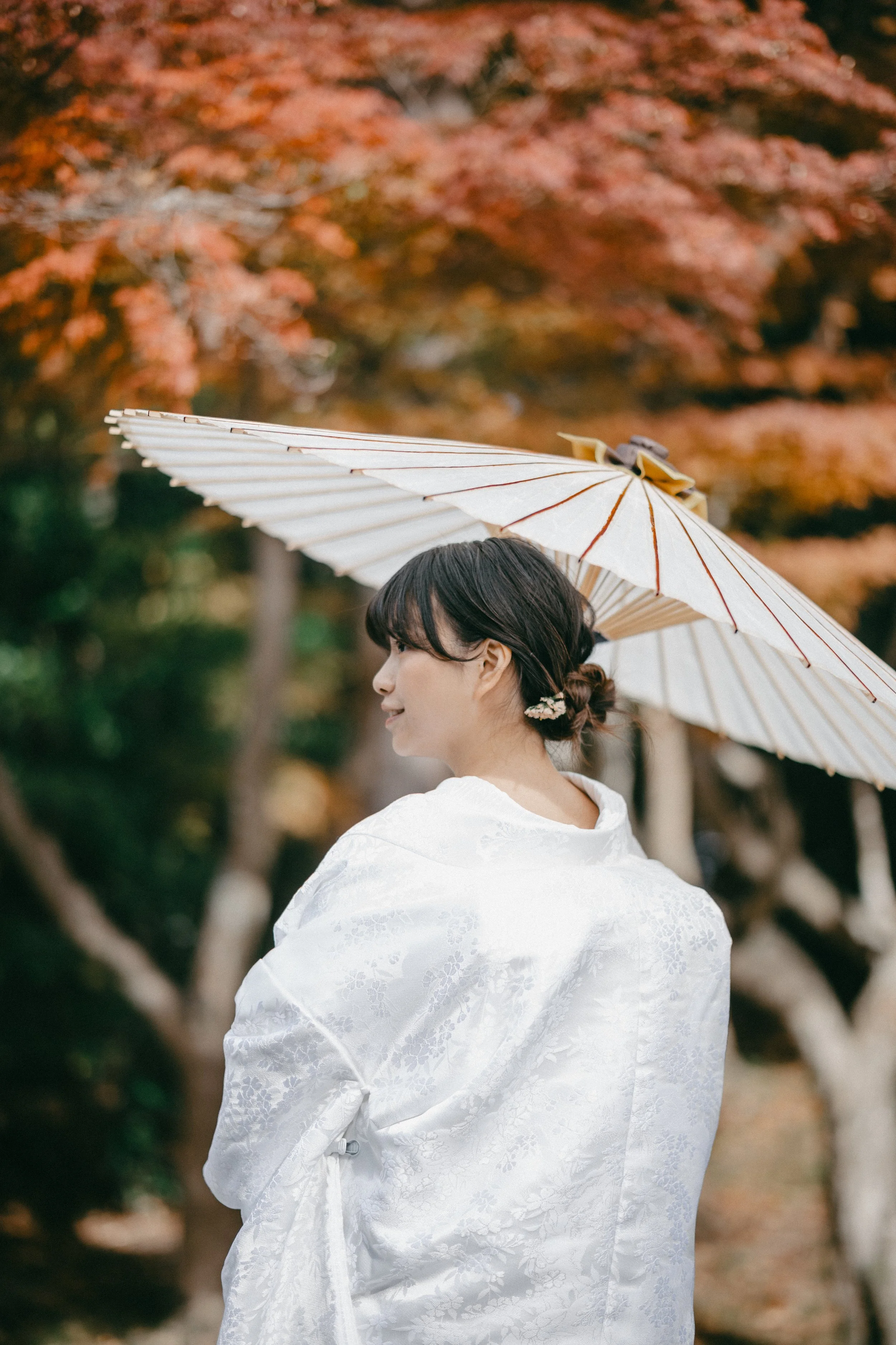 A woman in a white kimono holding a paper parasol with autumn trees in the background.