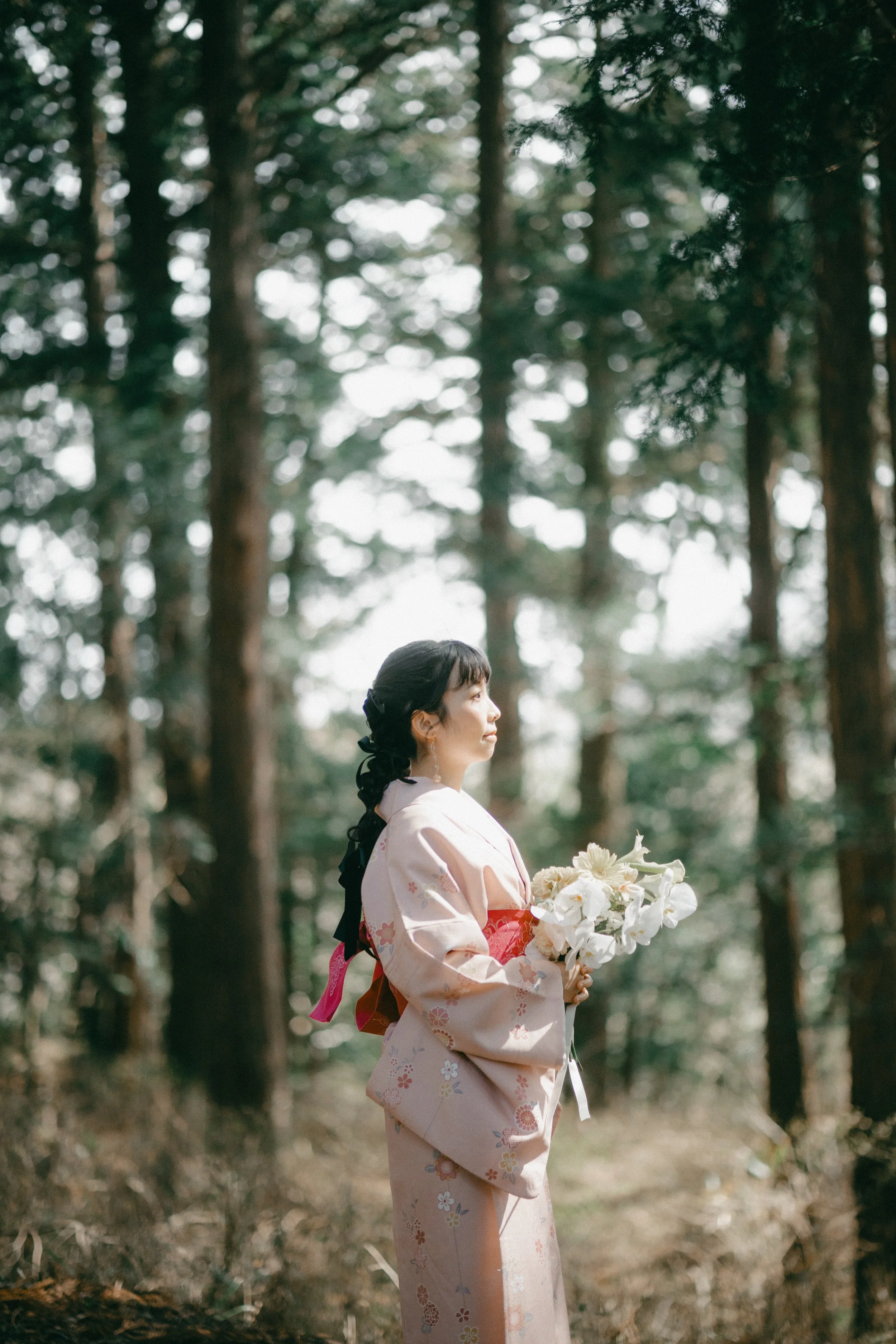A woman in a traditional kimono holding a bouquet of white flowers standing in a forest.