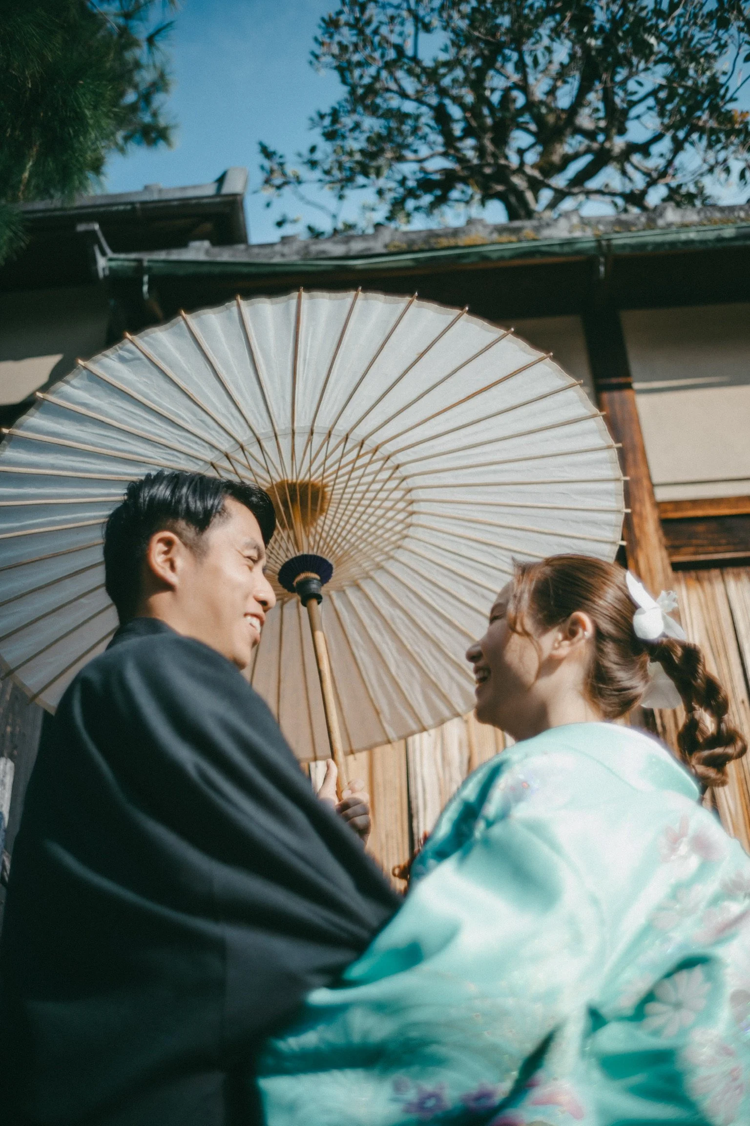 A smiling couple dressed in traditional Japanese attire holding a paper parasol outdoors against a background of a wooden building and a tree.