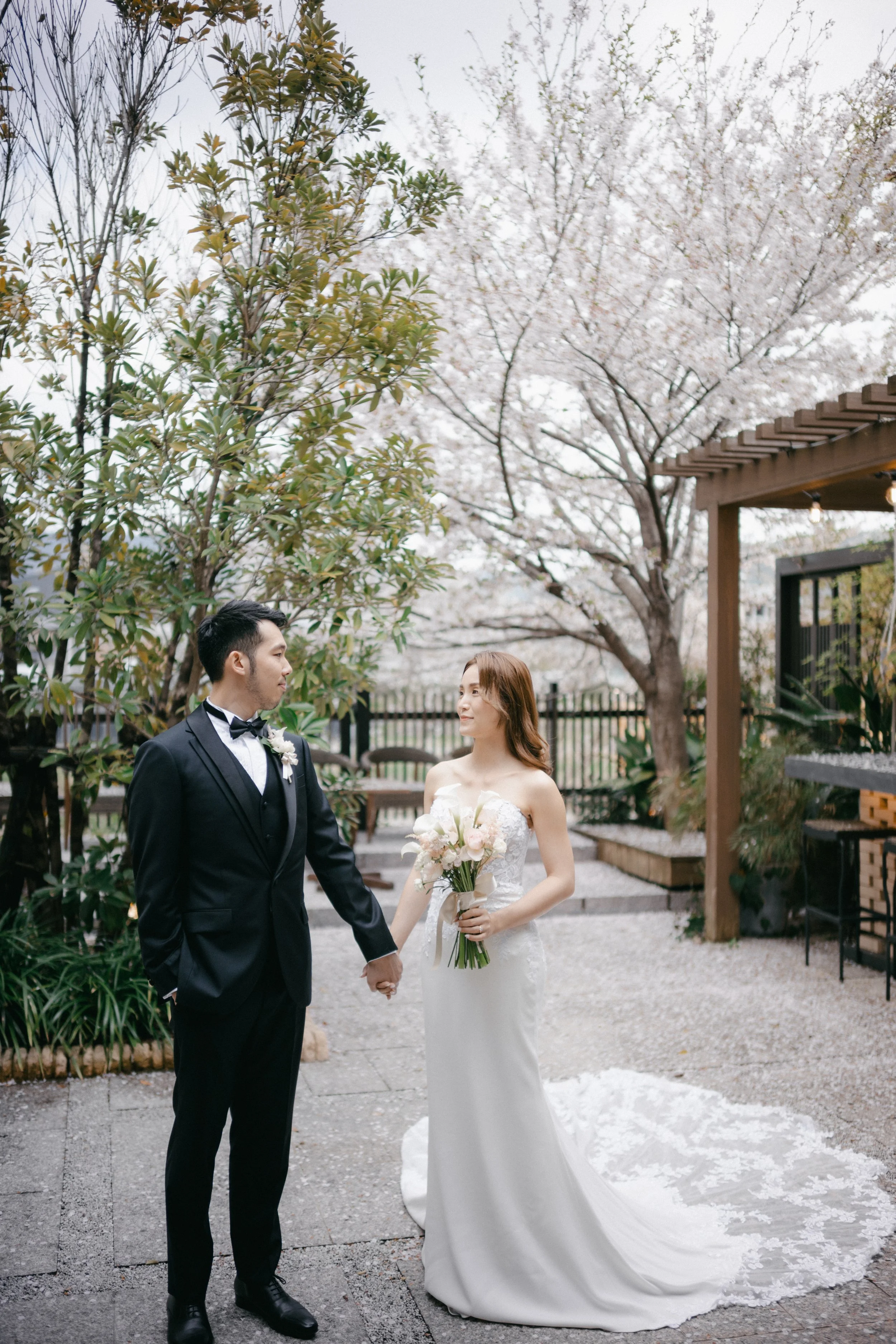 A bride and groom holding hands outdoors in a garden with blooming cherry blossom trees, the bride wearing a white strapless wedding gown and holding a bouquet, and the groom dressed in a black tuxedo with a bowtie.