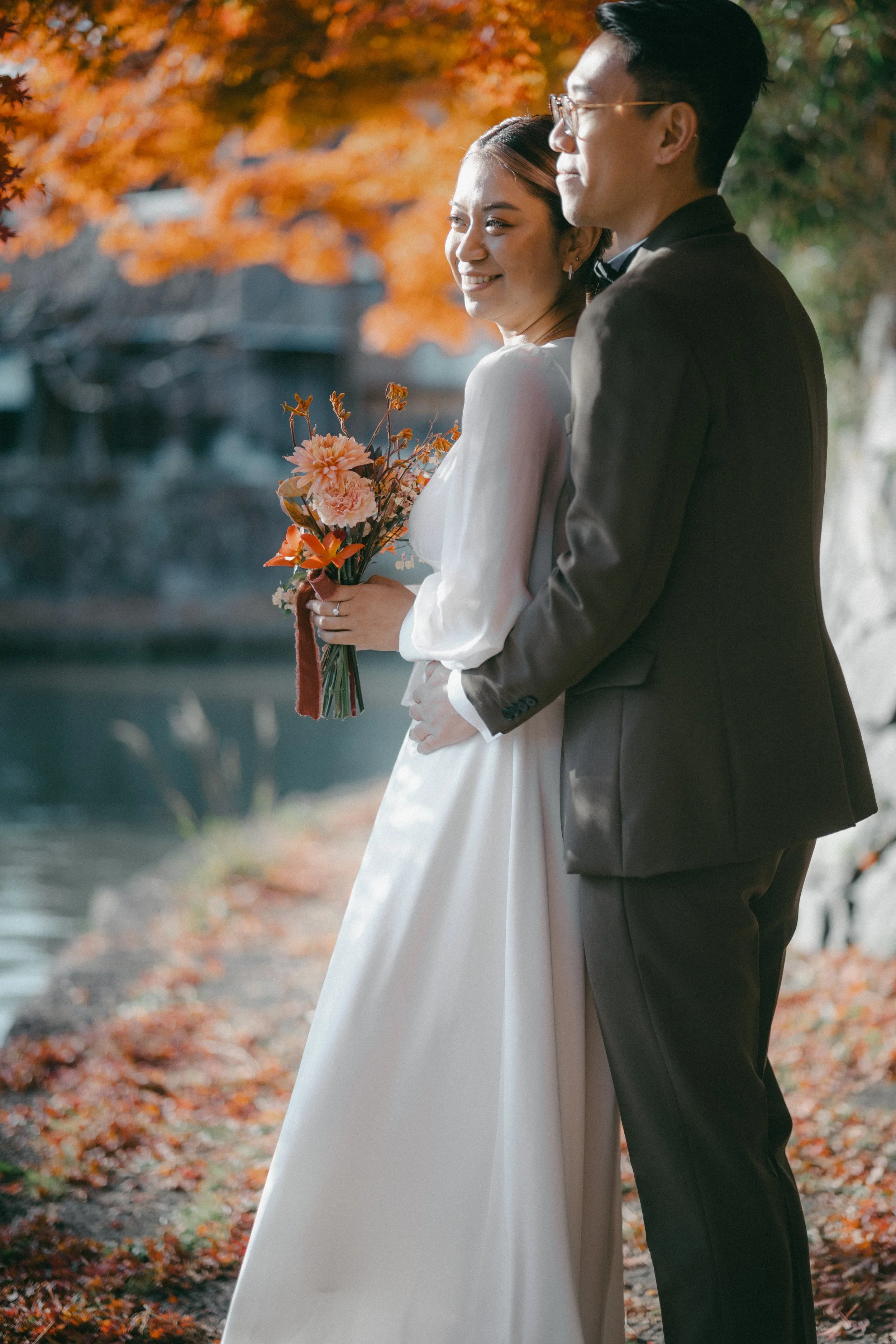 A bride and groom standing together outdoors, with autumn leaves in the background. The bride is holding a bouquet of pink and orange flowers, and both are smiling.