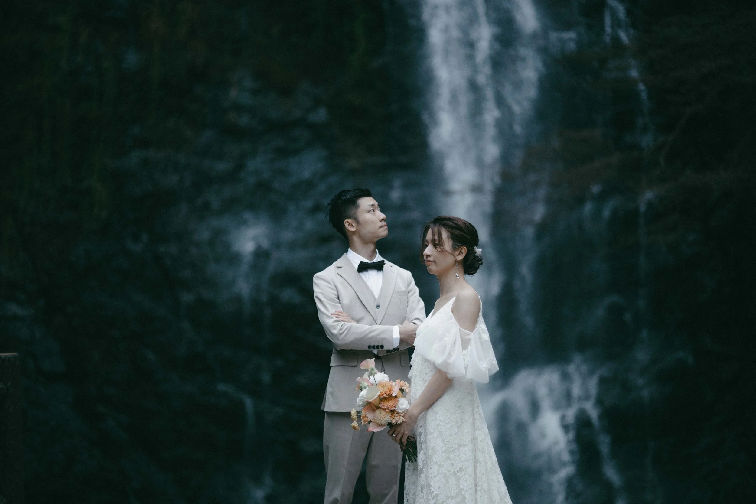 A couple dressed in wedding attire standing in front of a waterfall, the groom with arms crossed wearing a beige suit and bowtie, and the bride holding a bouquet of flowers, gazing at each other.