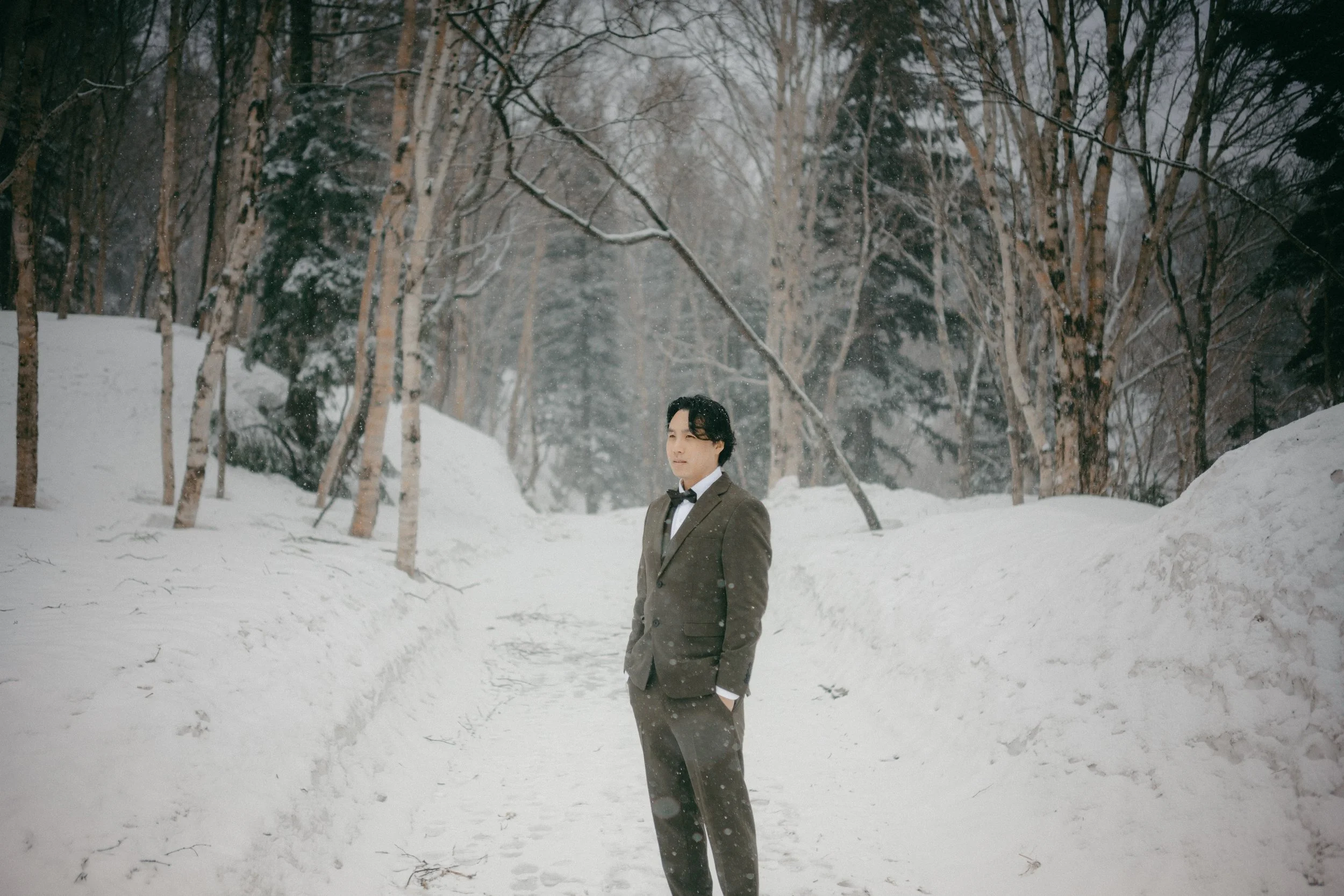 A man in a suit and bow tie standing in a snowy forest with snow-covered ground and leafless trees.