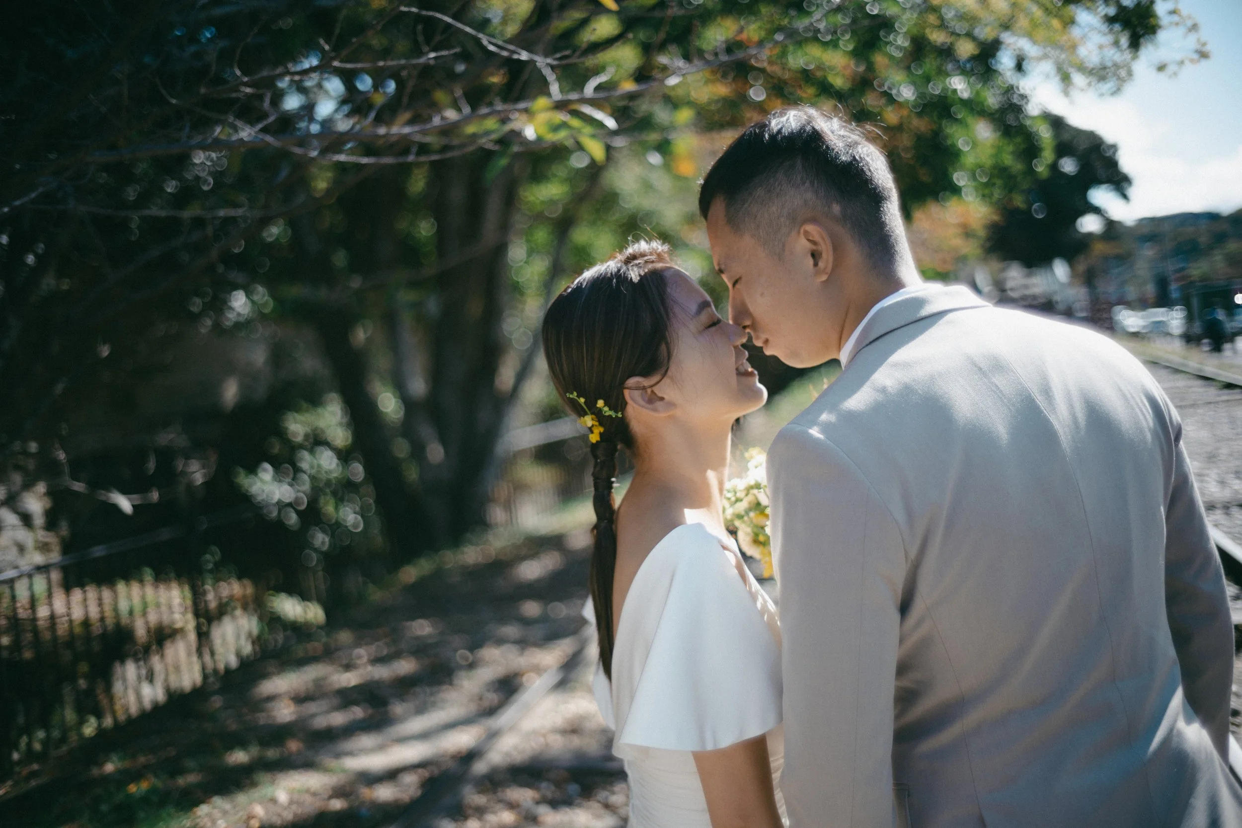 A newlywed couple sharing an intimate moment outdoors on a sunny day, with trees and a train track in the background.