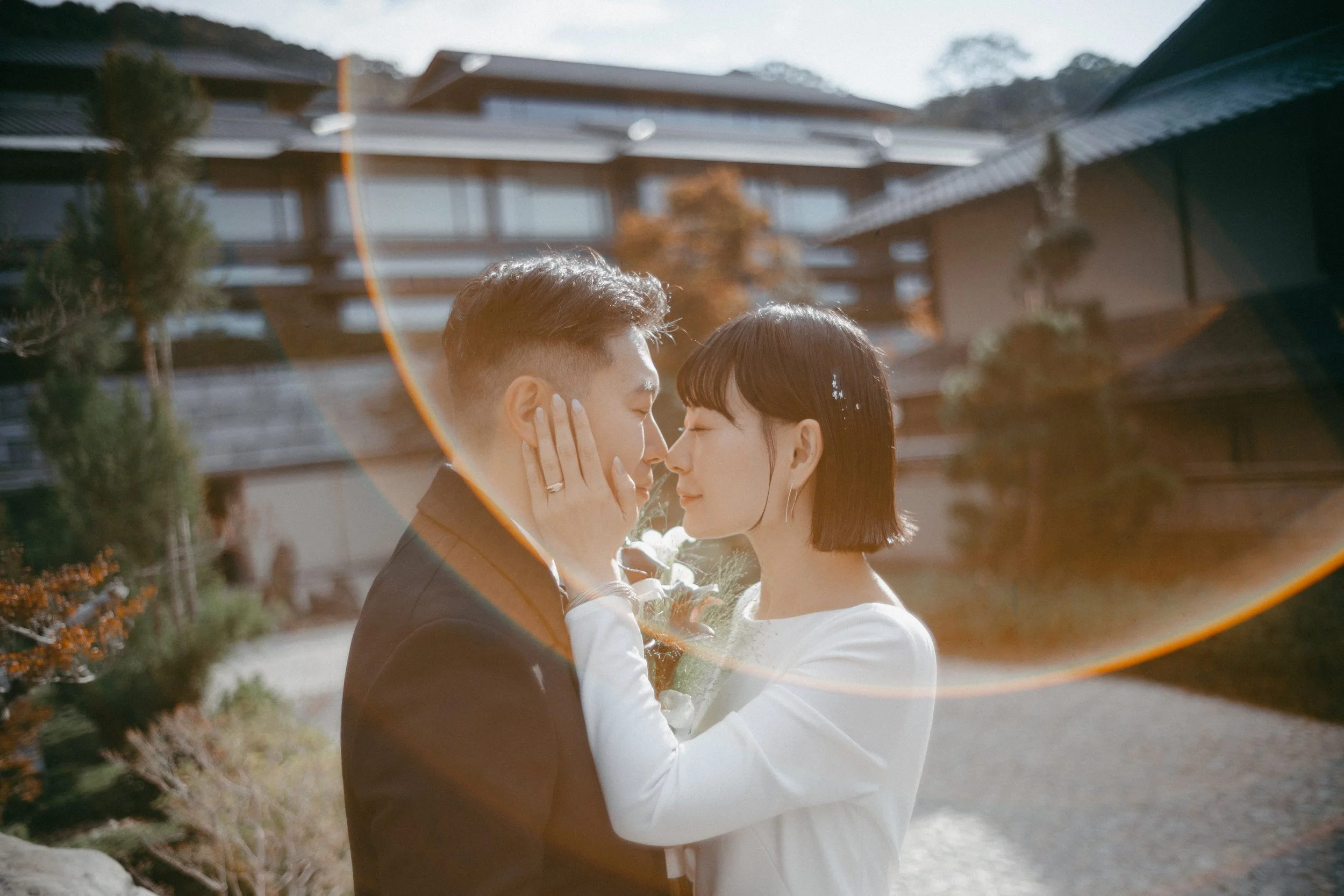 A couple standing close together outside, touching foreheads and smiling, with sun flare and mountains in the background.
