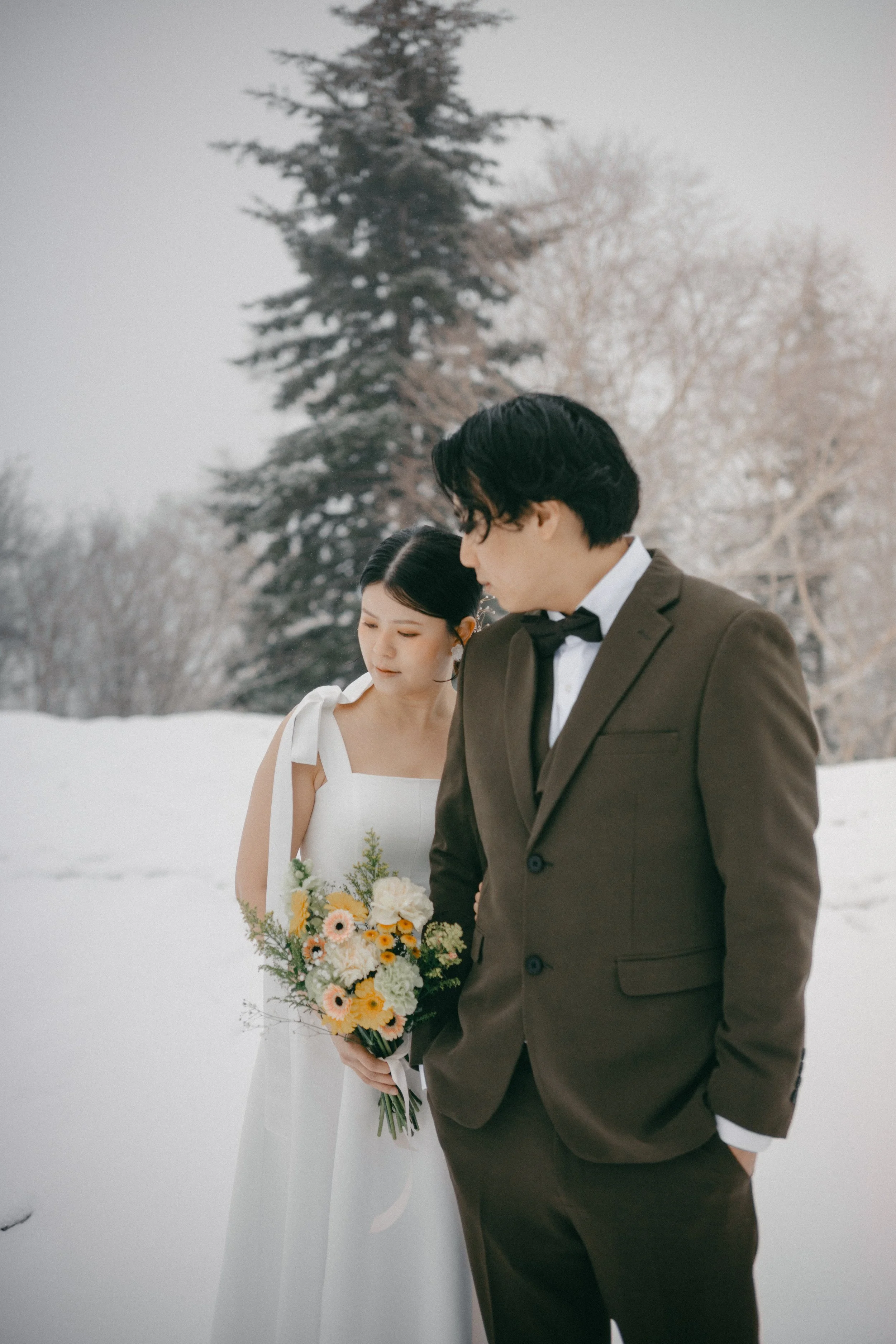 A bride and groom stand close together outdoors in a snowy landscape with trees in the background, the bride holding a colorful bouquet of flowers.