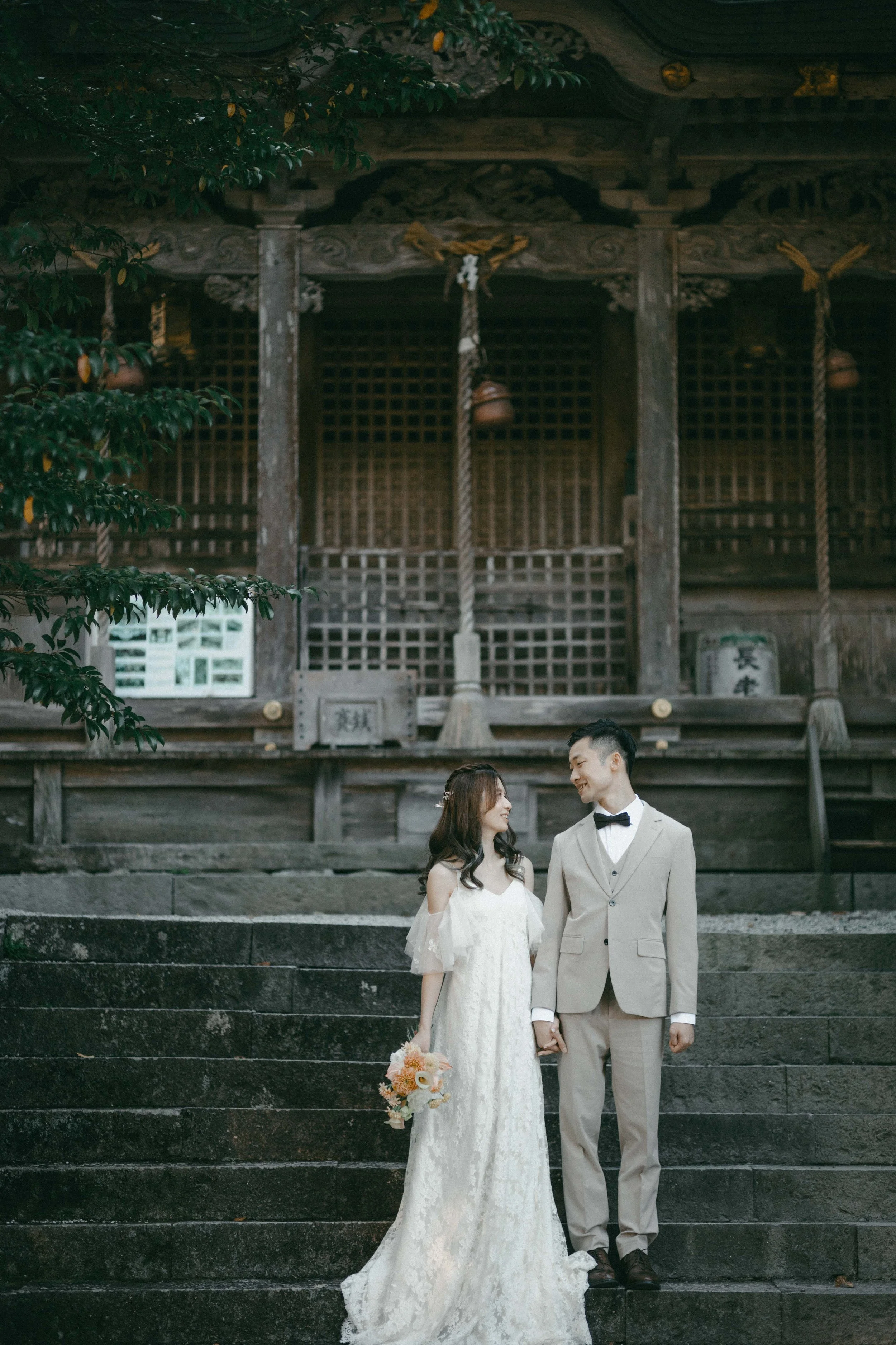 A bride and groom holding hands, standing on stone steps outside a traditional wooden temple, dressed in wedding attire, with the bride holding a bouquet of flowers, smiling at each other.