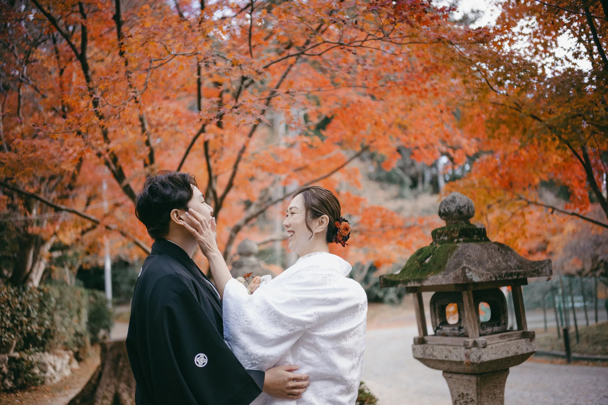 A couple dressed in traditional Japanese wedding attire standing close together outdoors during autumn, surrounded by orange and red maple trees and a stone lantern.