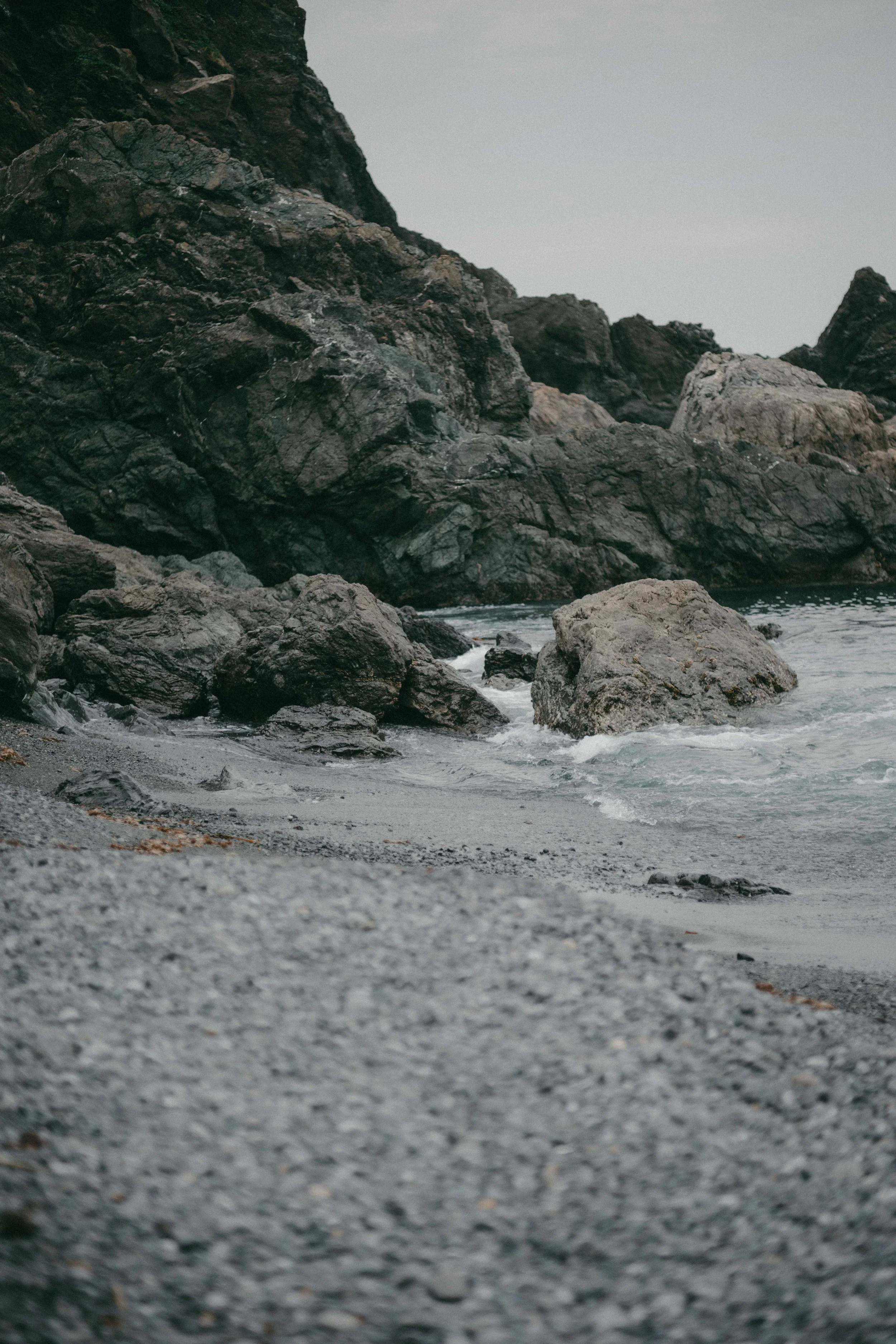 A rocky beach with large dark rocks along the shore and water gently washing over smaller pebbles.