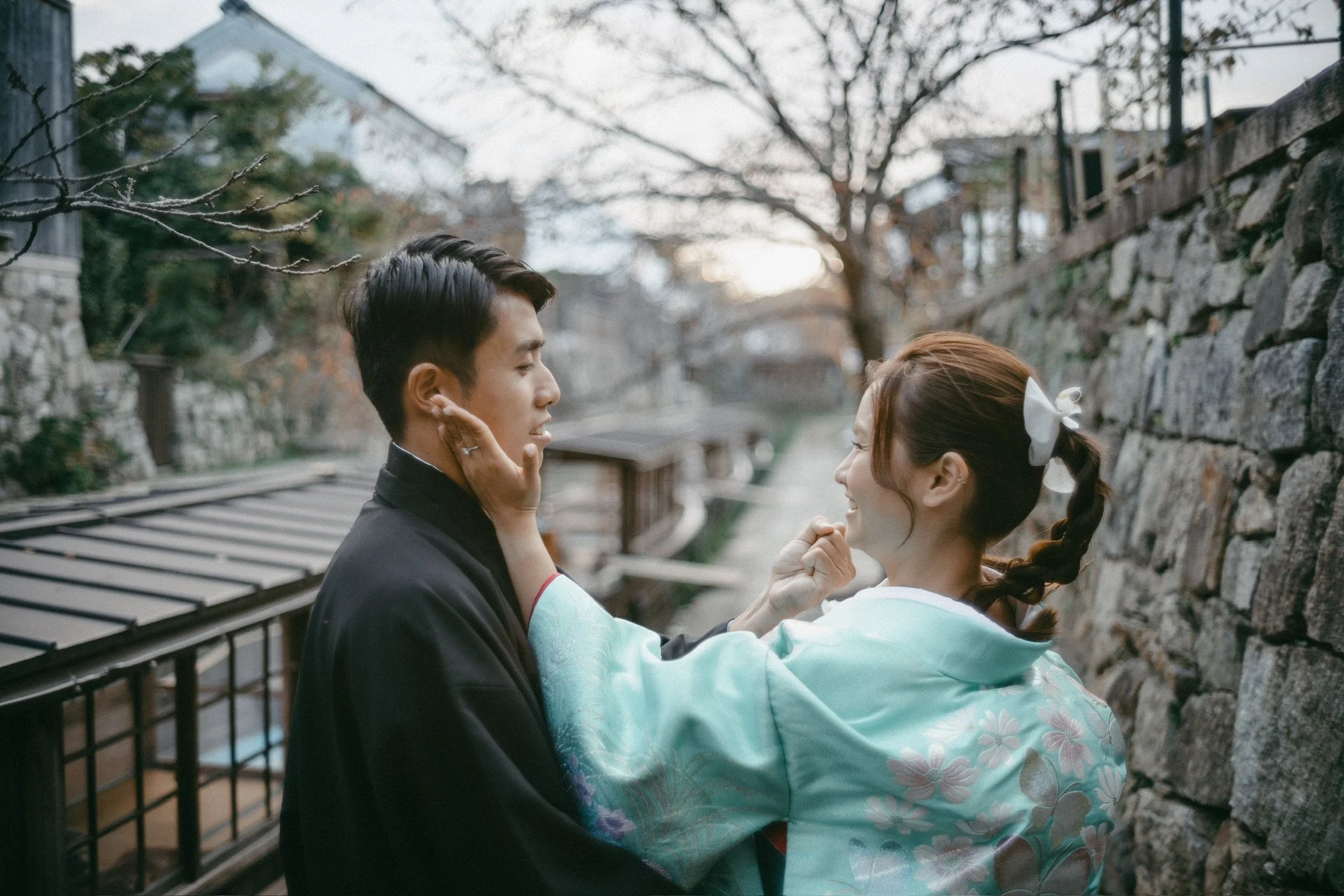 A man and woman face each other outdoors, with the woman gently touching the man's face. The woman is smiling, and the scene is set against a stone wall and trees without leaves.