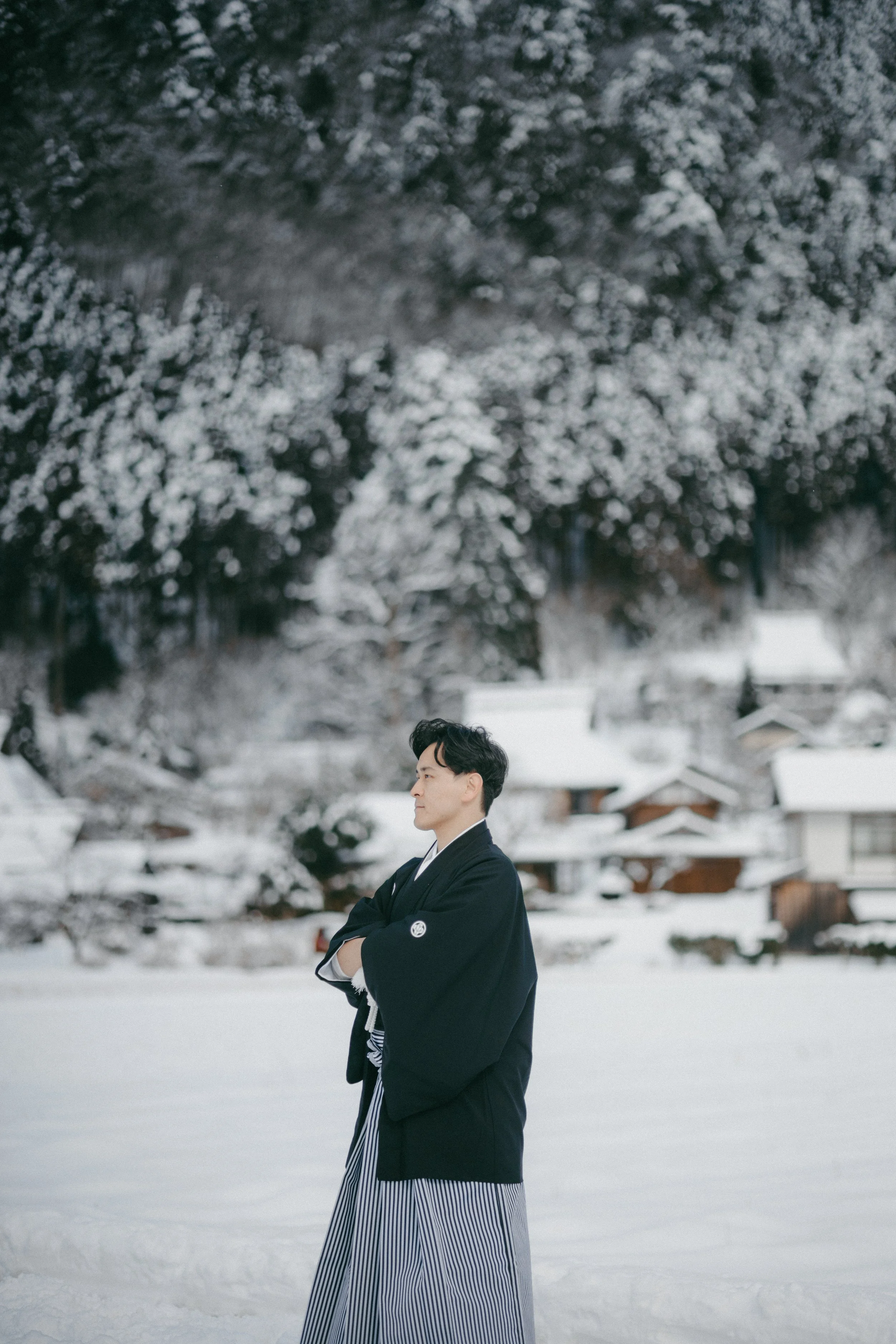 A man dressed in traditional Japanese clothing standing in a snowy landscape with houses and snow-covered trees in the background.