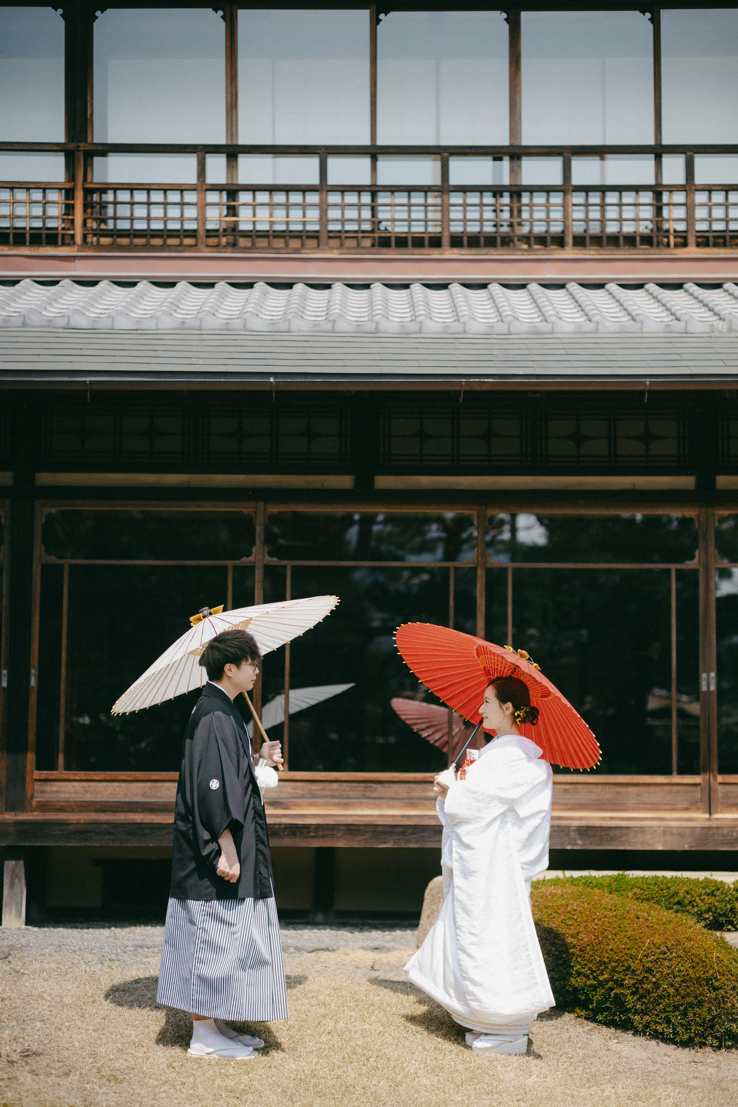 A bride and groom in traditional Japanese wedding attire holding umbrellas and facing each other outside a wooden building.
