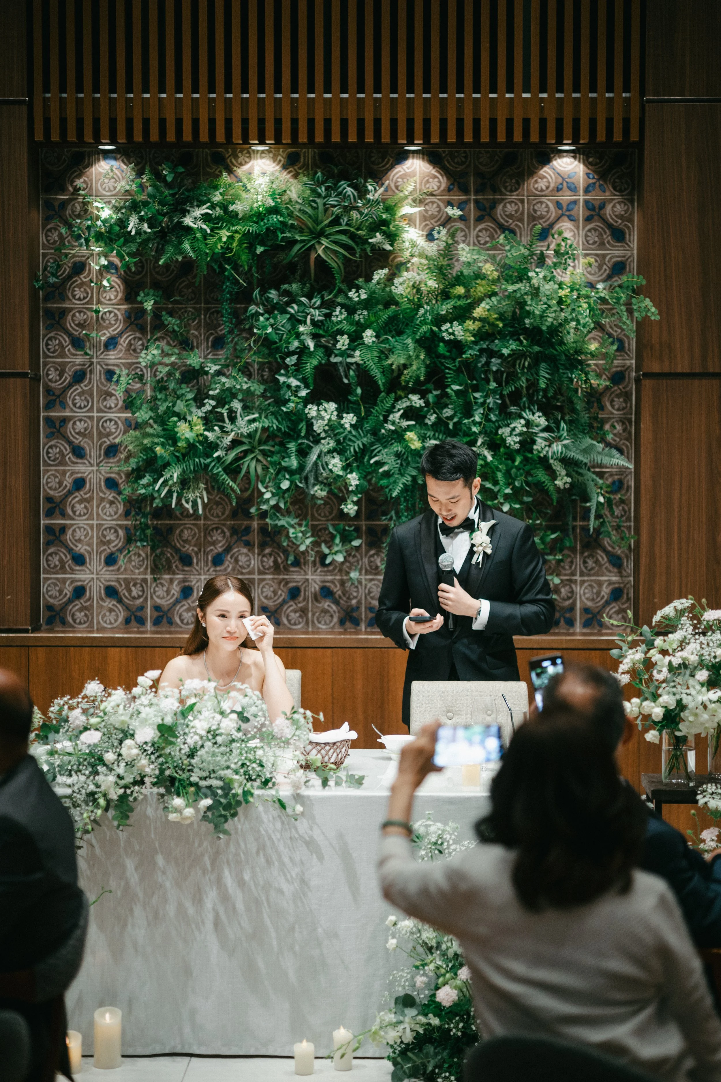 A wedding reception with a bride and groom seated at a decorated table, the groom giving a speech with a microphone, surrounded by guests, floral arrangements, and candles.