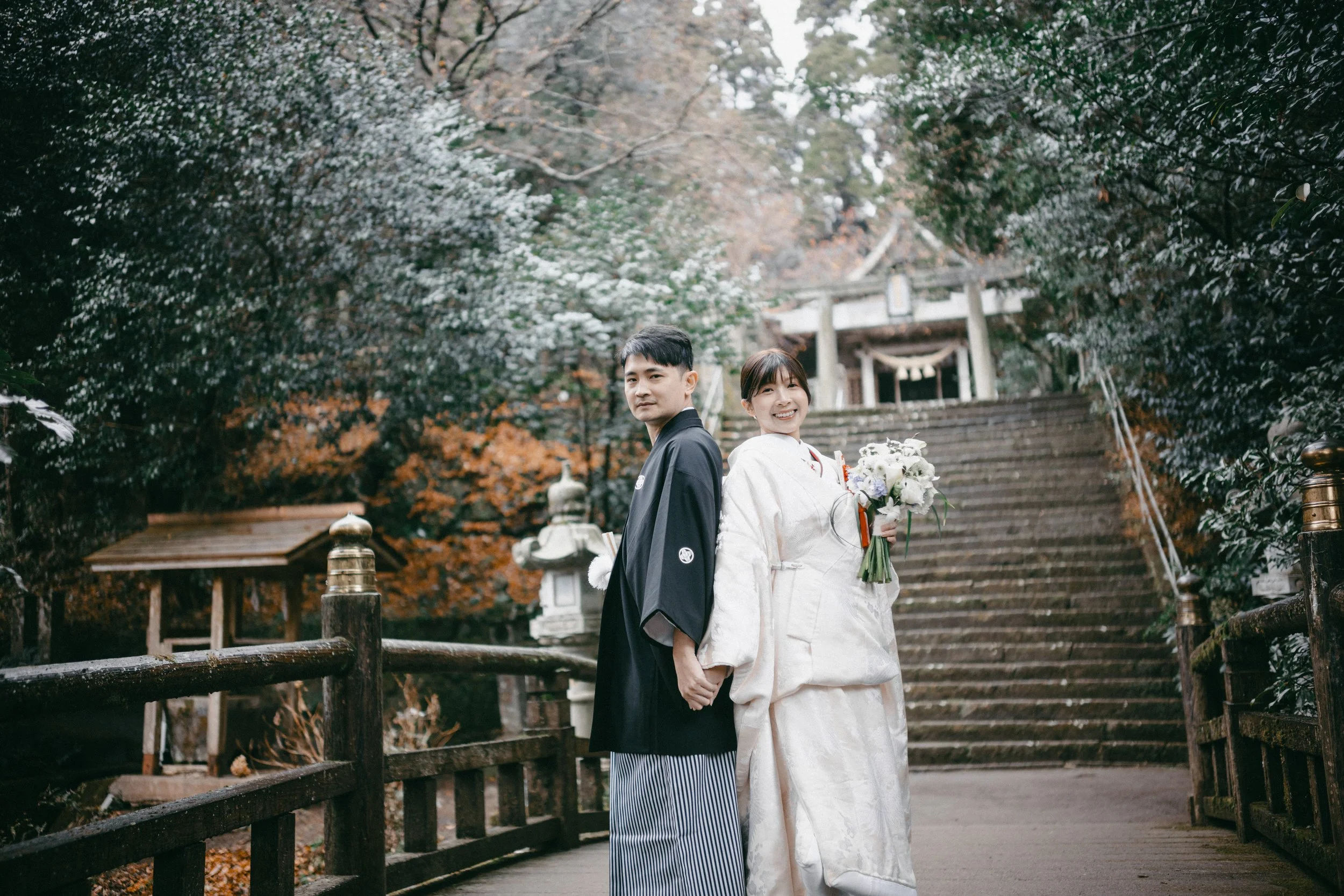 A newlywed couple in traditional Japanese wedding attire holding hands and smiling, standing on steps at a shrine surrounded by autumn foliage.