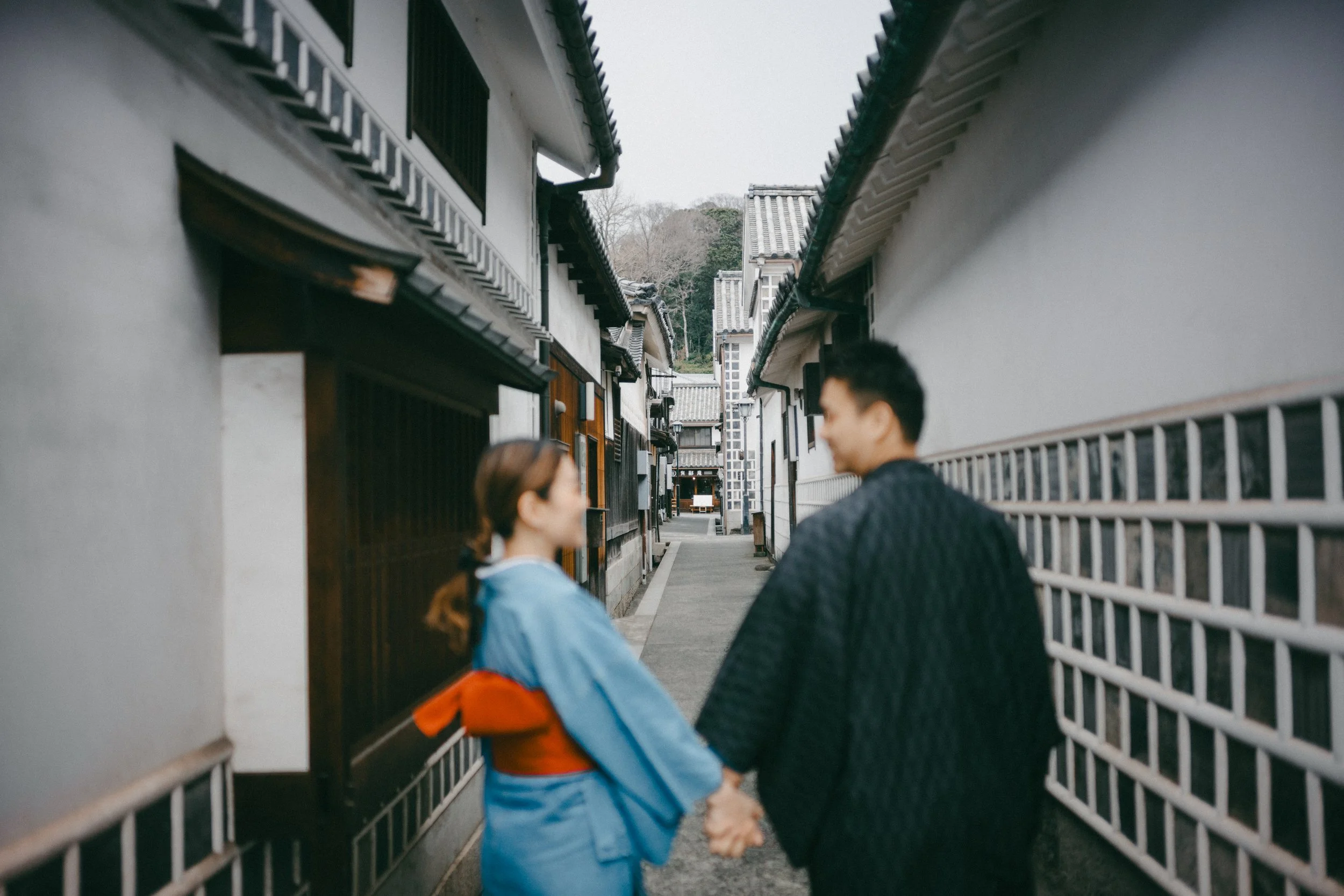A couple holding hands and smiling at each other walking through a narrow alley with traditional Japanese houses.