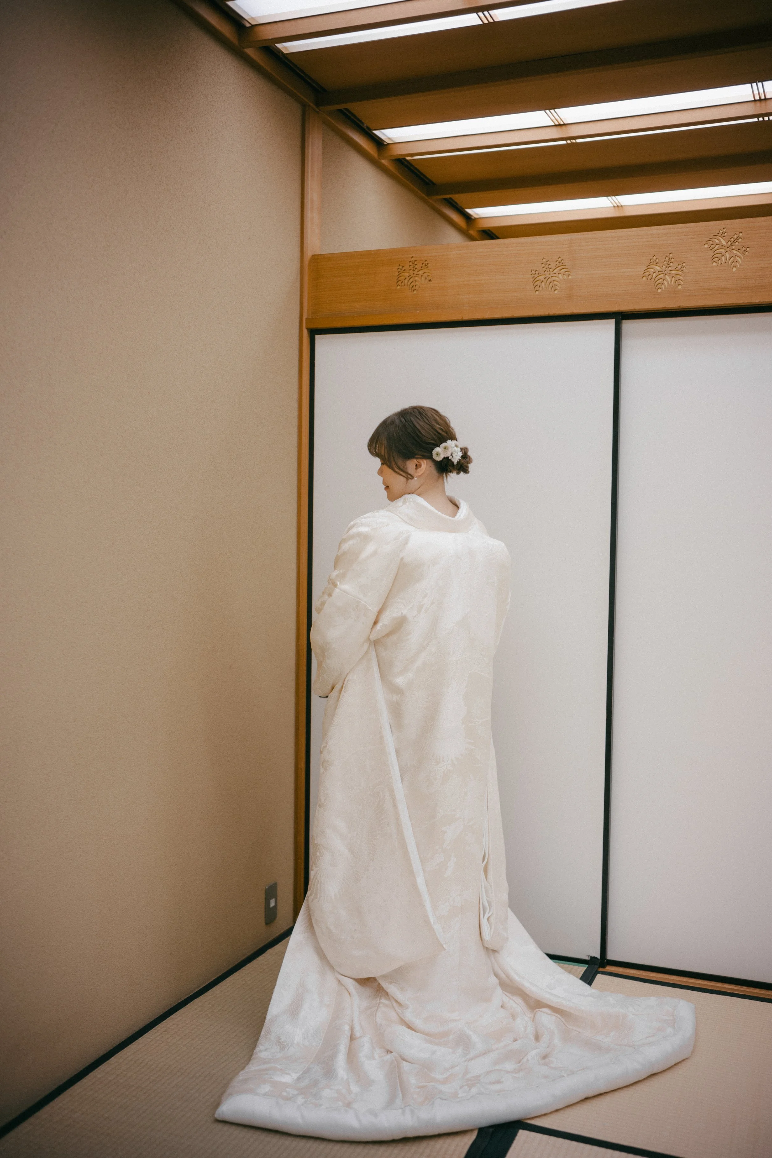 A woman in a traditional Japanese wedding kimono, with a floral hair accessory, standing in a tatami room with wooden accents.
