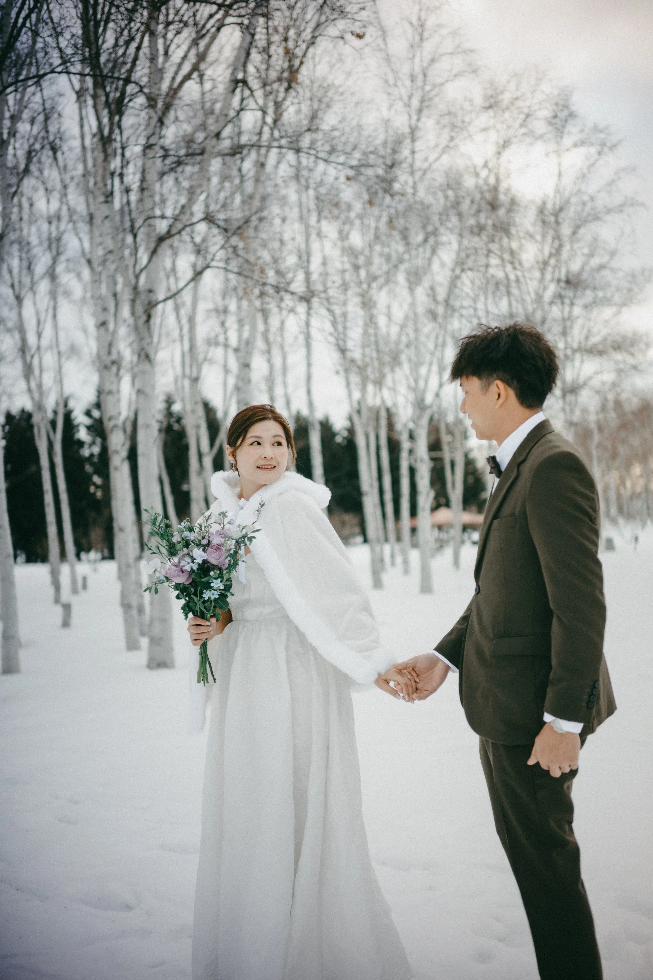 A bride and groom holding hands in a snowy forest, the bride holding a bouquet of purple, white, and green flowers.