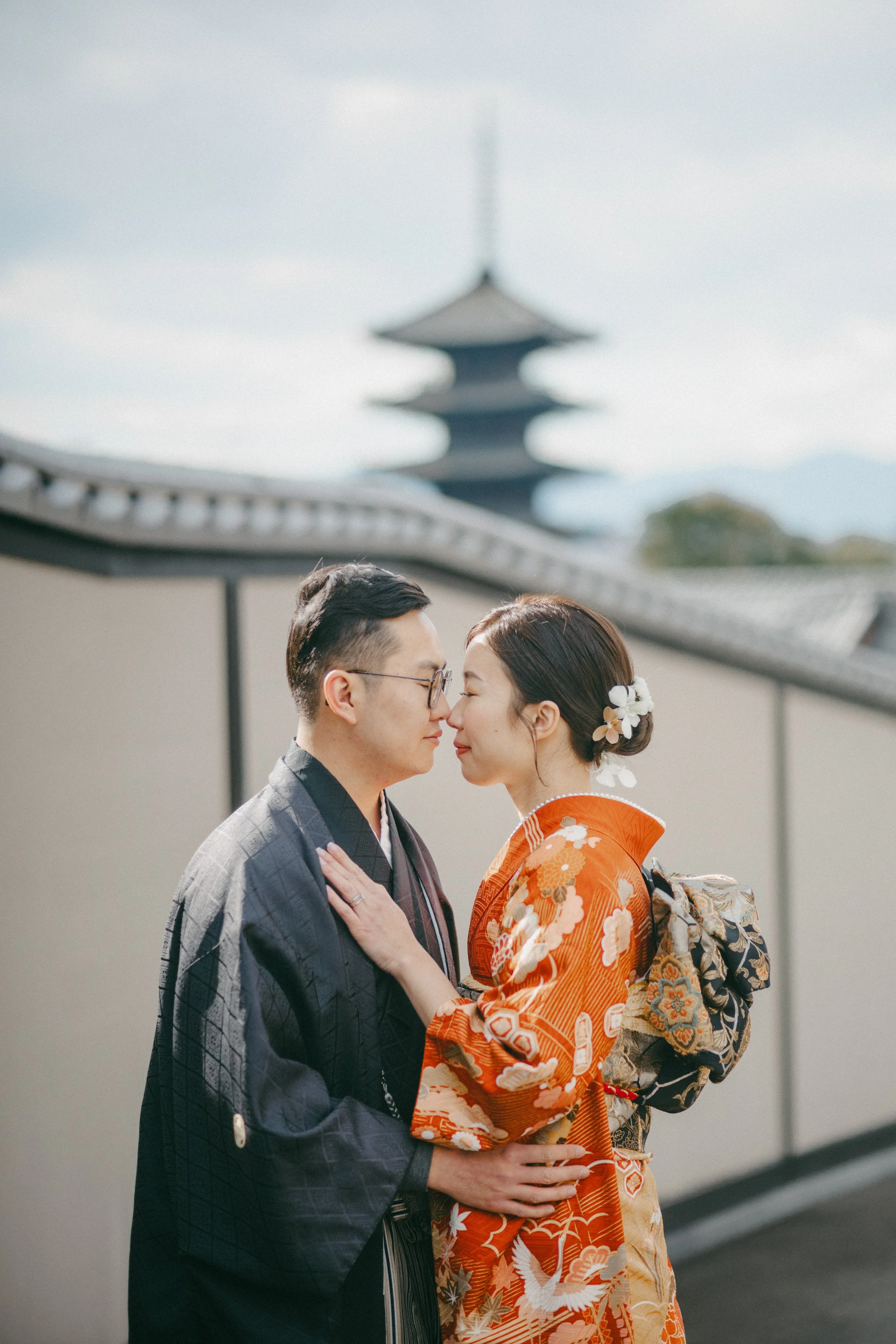 A couple dressed in traditional Japanese kimonos in an outdoor setting with a pagoda in the background, sharing a close moment with foreheads touching.