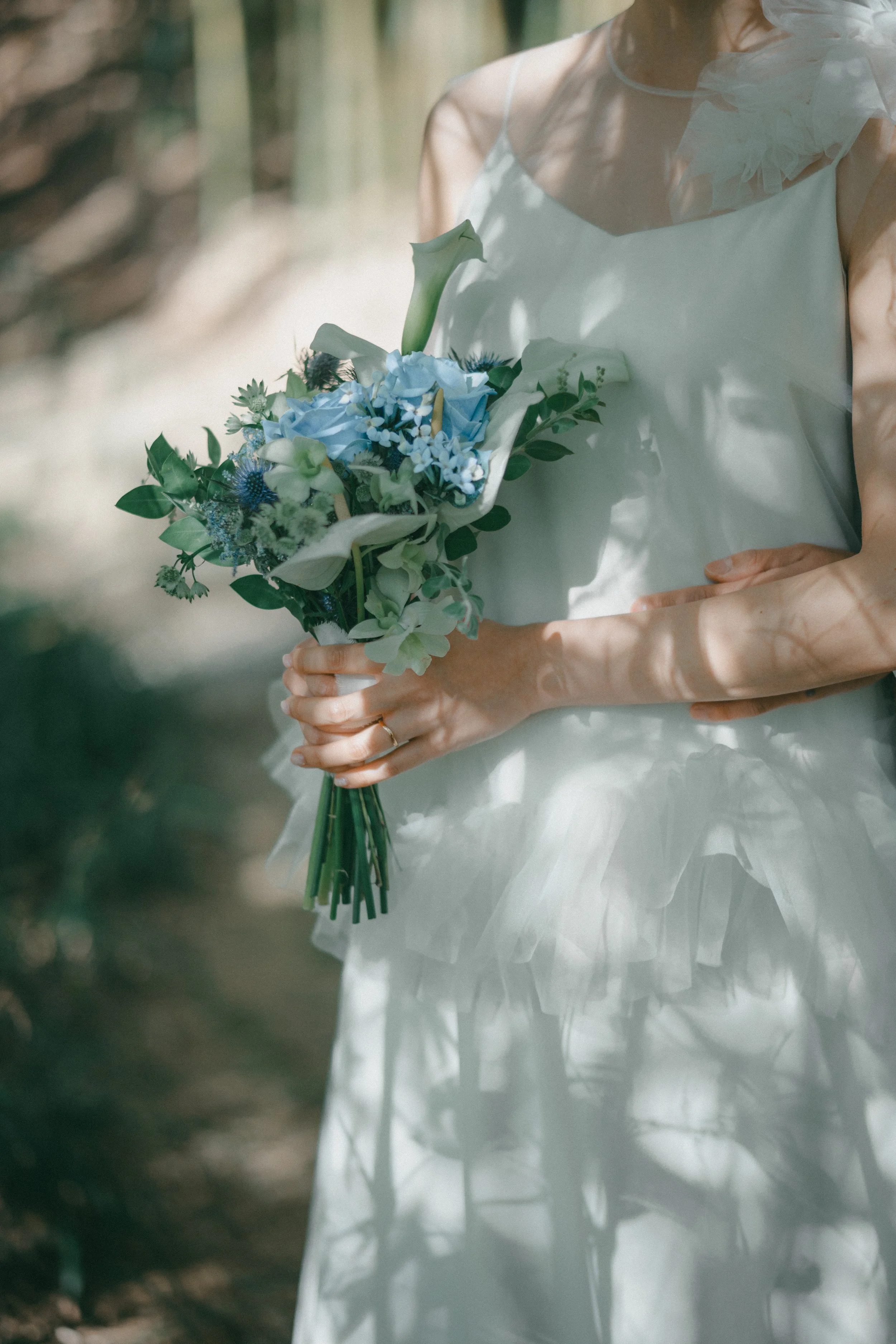 A woman in a white dress holding a bouquet of white and blue flowers in an outdoor setting with sunlight and shadows.