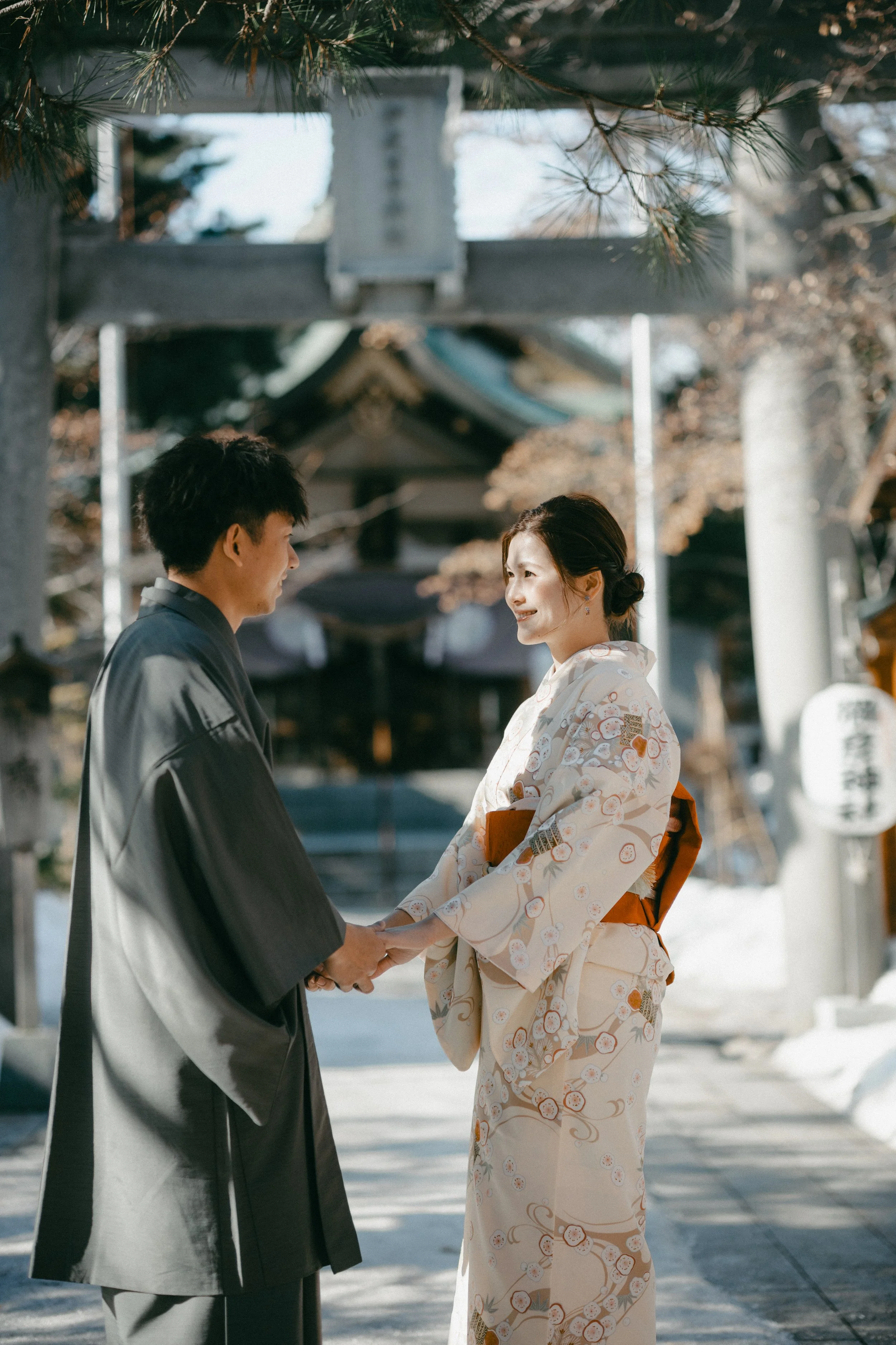 A Japanese couple holding hands and looking at each other, standing outdoors in a traditional setting with a shrine or temple in the background.