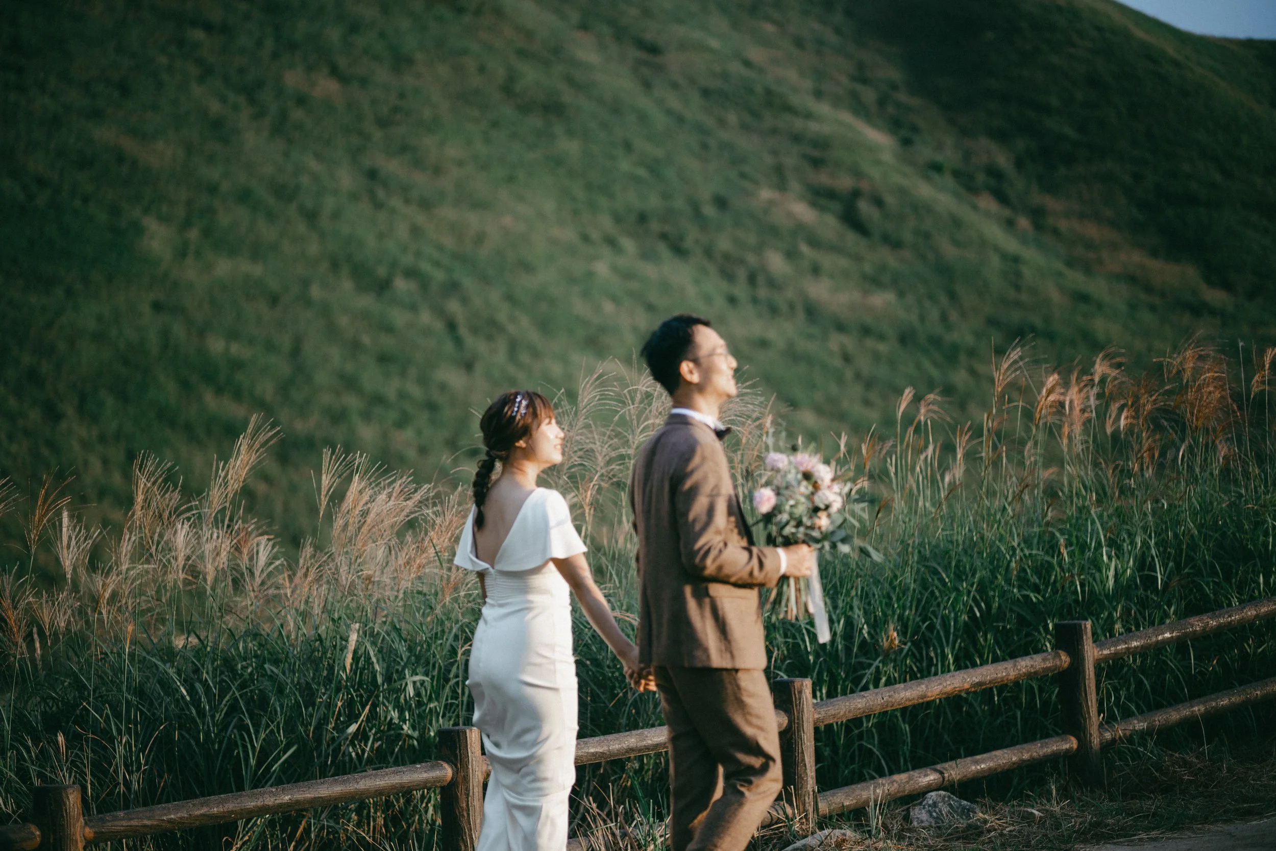 A couple dressed in wedding attire walking hand in hand along a trail next to tall grass fields with a green hillside in the background.