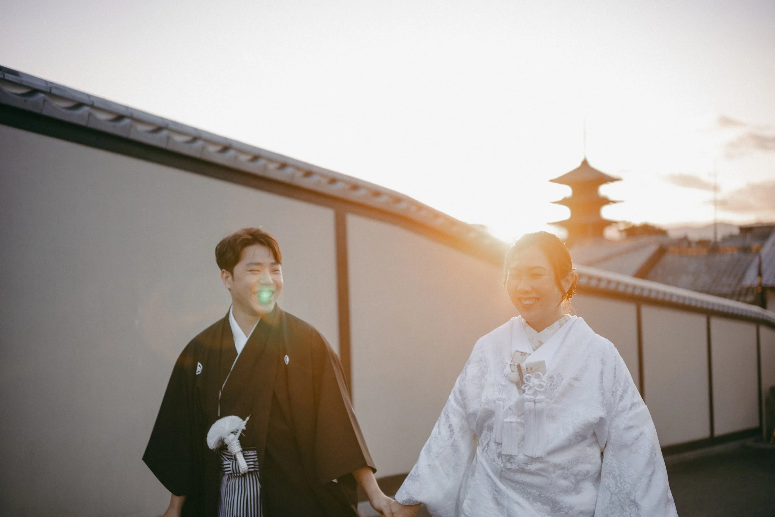 A couple dressed in traditional Japanese wedding attire, holding hands and smiling at each other during sunset with a temple in the background.