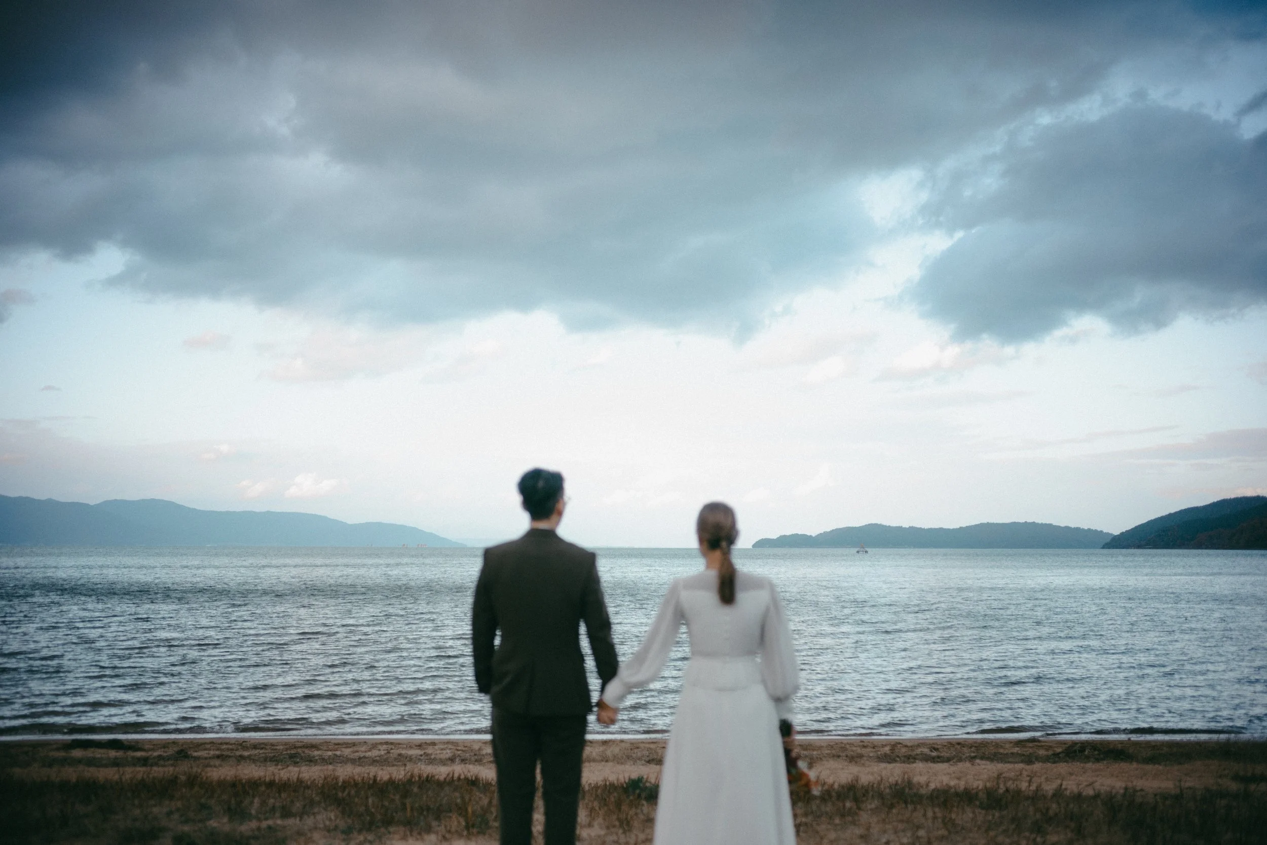 A man and woman in wedding attire standing hand-in-hand on a beach, looking out over a calm body of water with distant islands and dark, cloudy sky