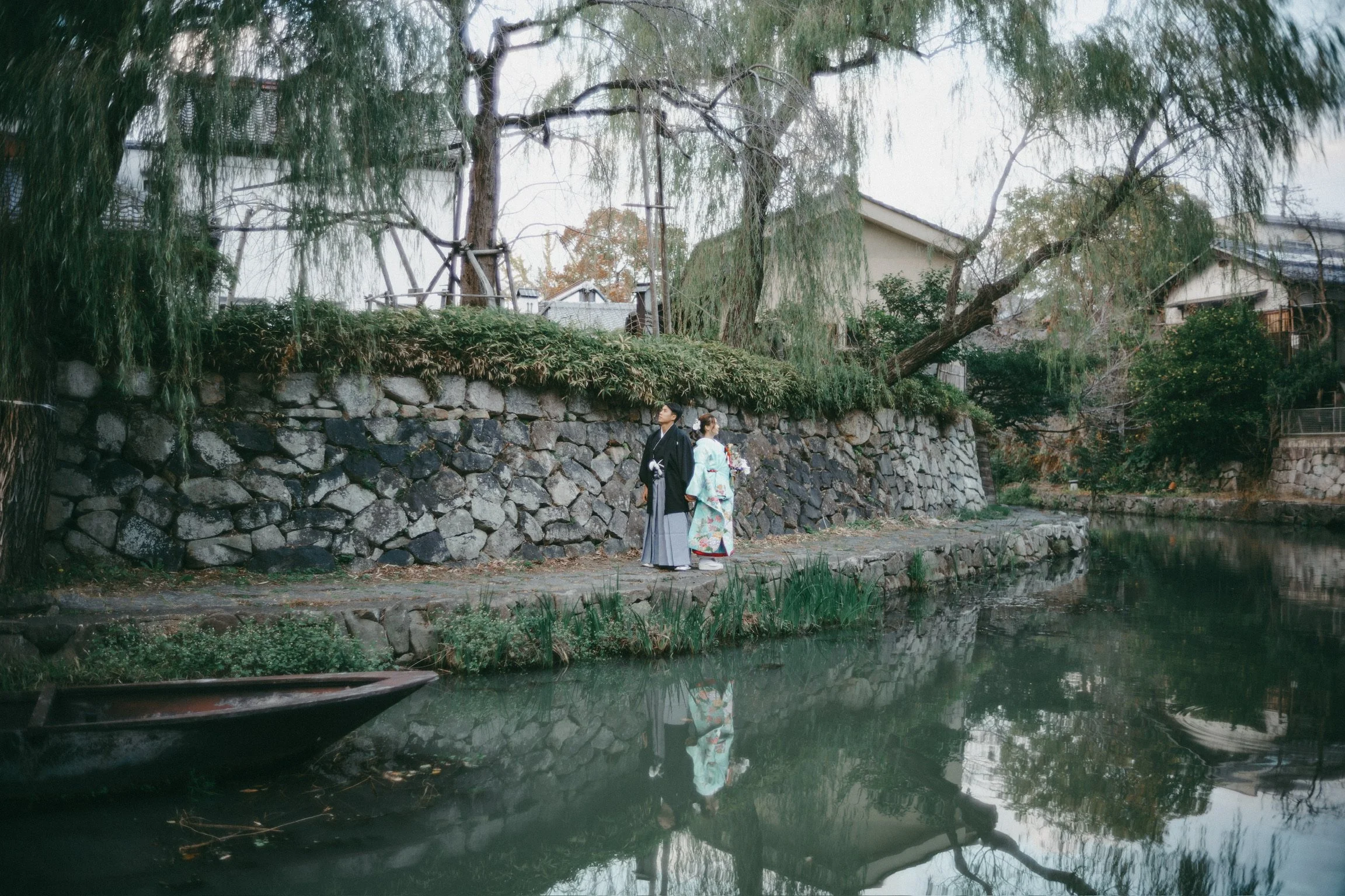 A couple dressed in traditional Japanese kimonos stand by a canal, with lush green trees and stone walls in the background.