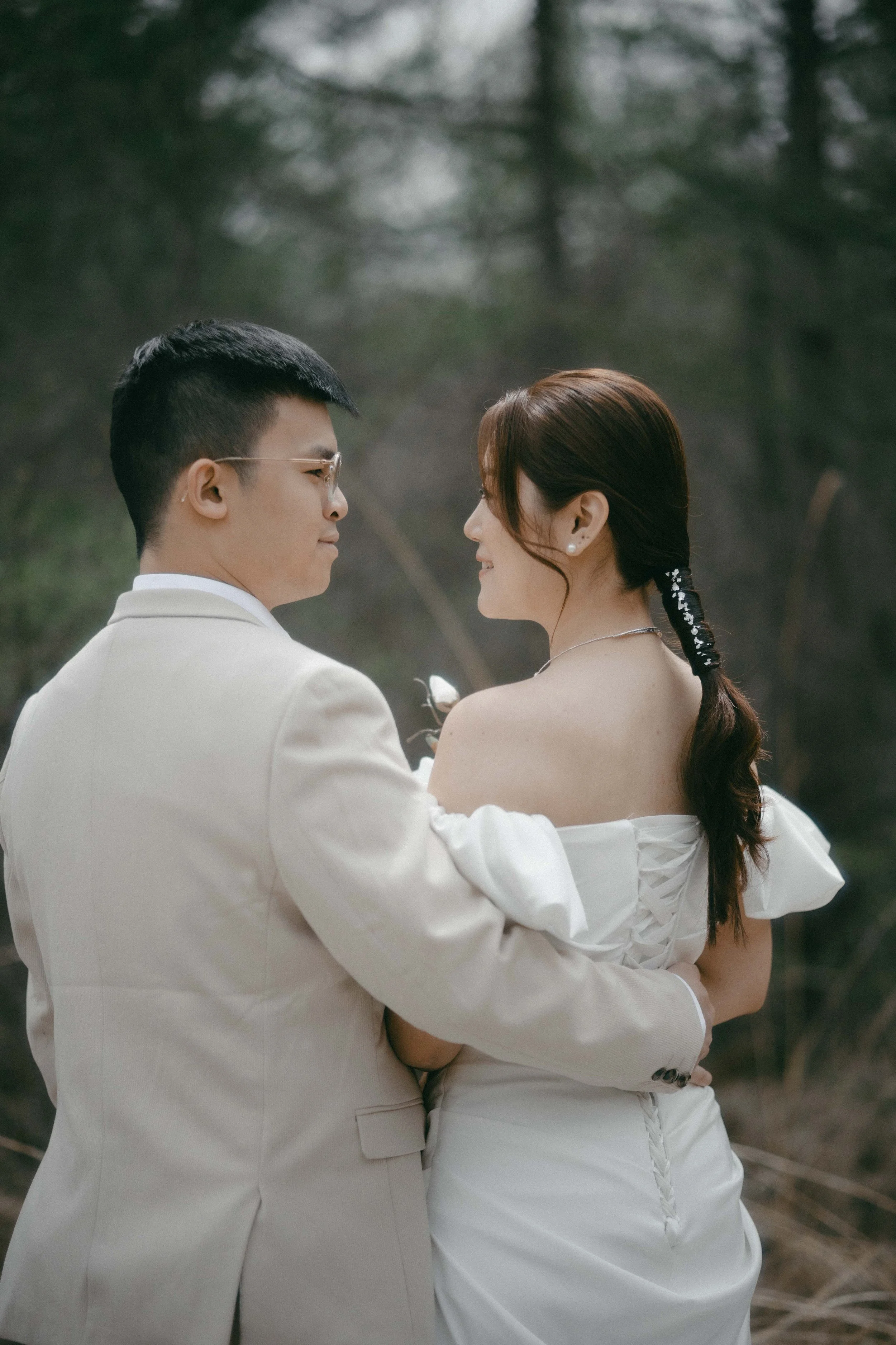 A couple in wedding attire embracing outdoors with trees in the background.
