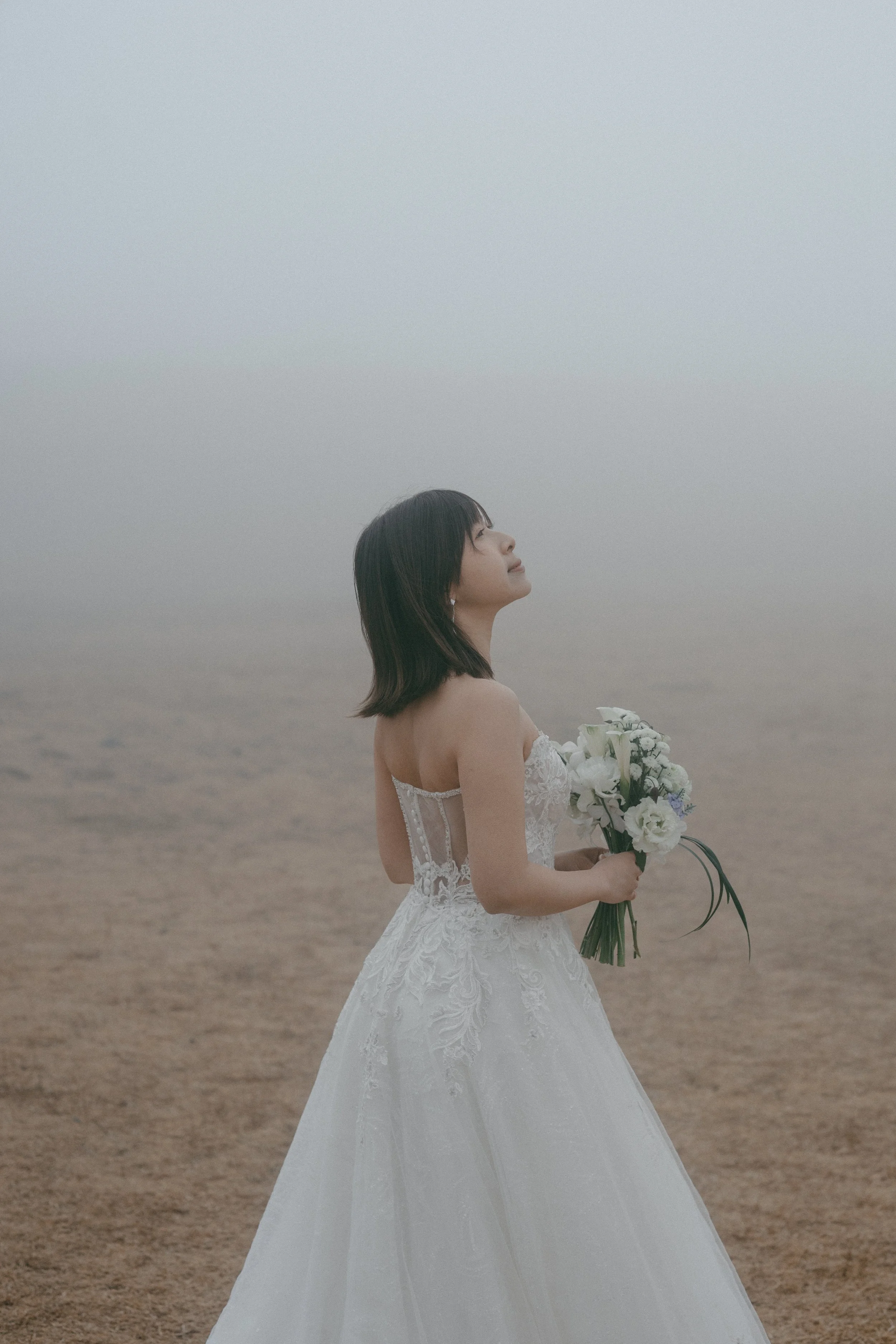 A woman in a wedding dress holding a bouquet, standing on a foggy beach.