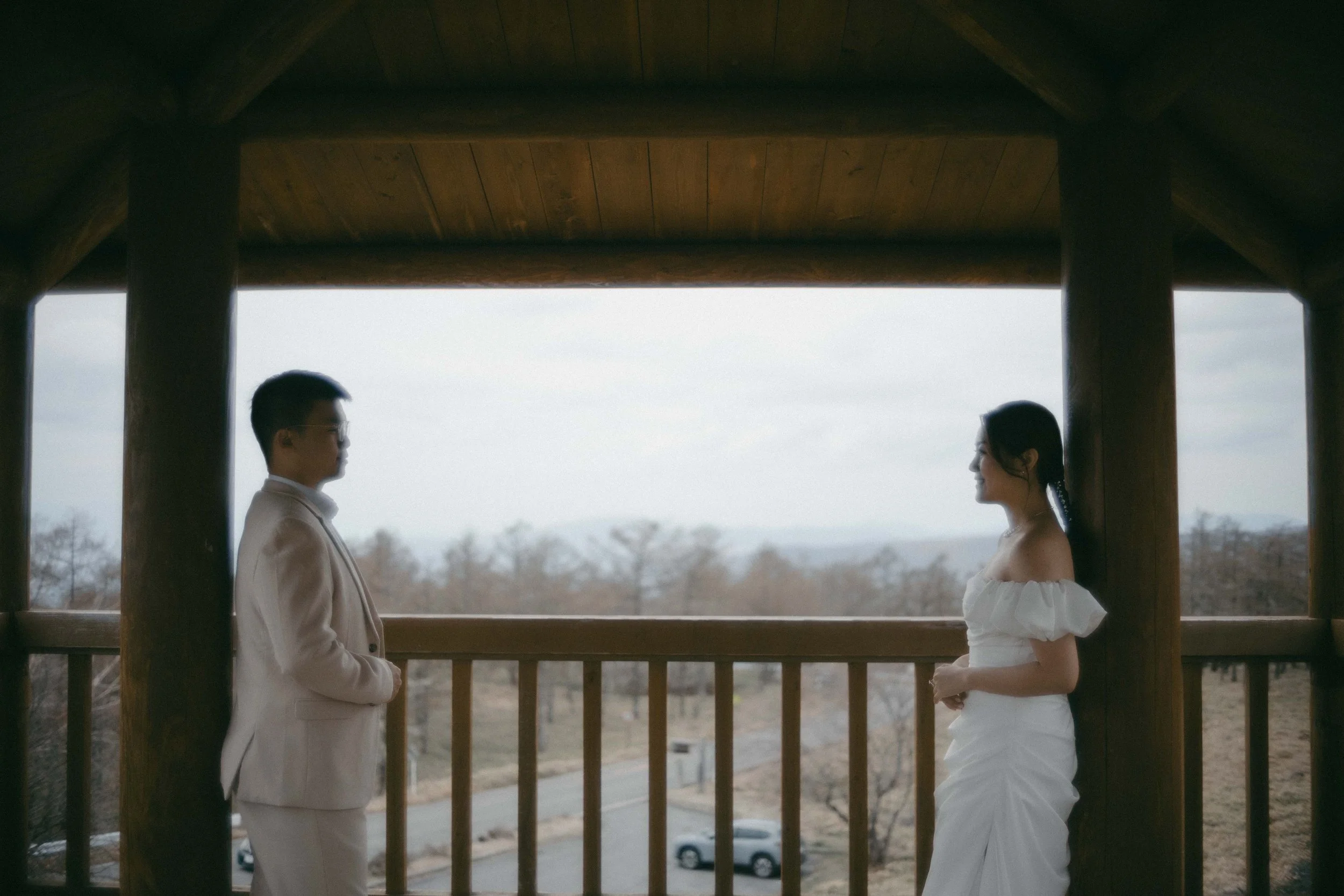 A man and woman face each other on a wooden balcony, with a scenic outdoor landscape in the background.